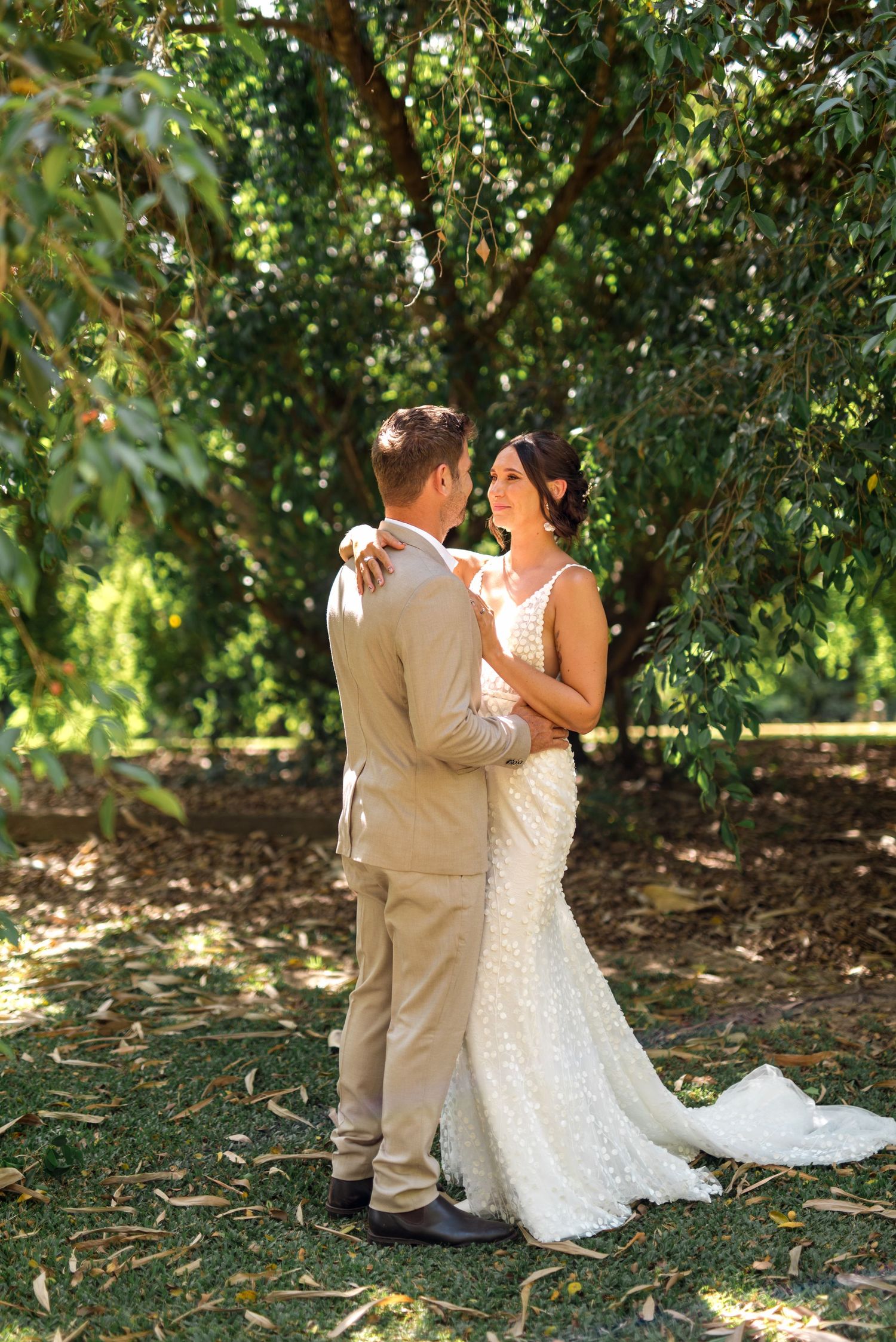 Bride and groom embrace tenderly under lush green trees in an outdoor wedding portrait.