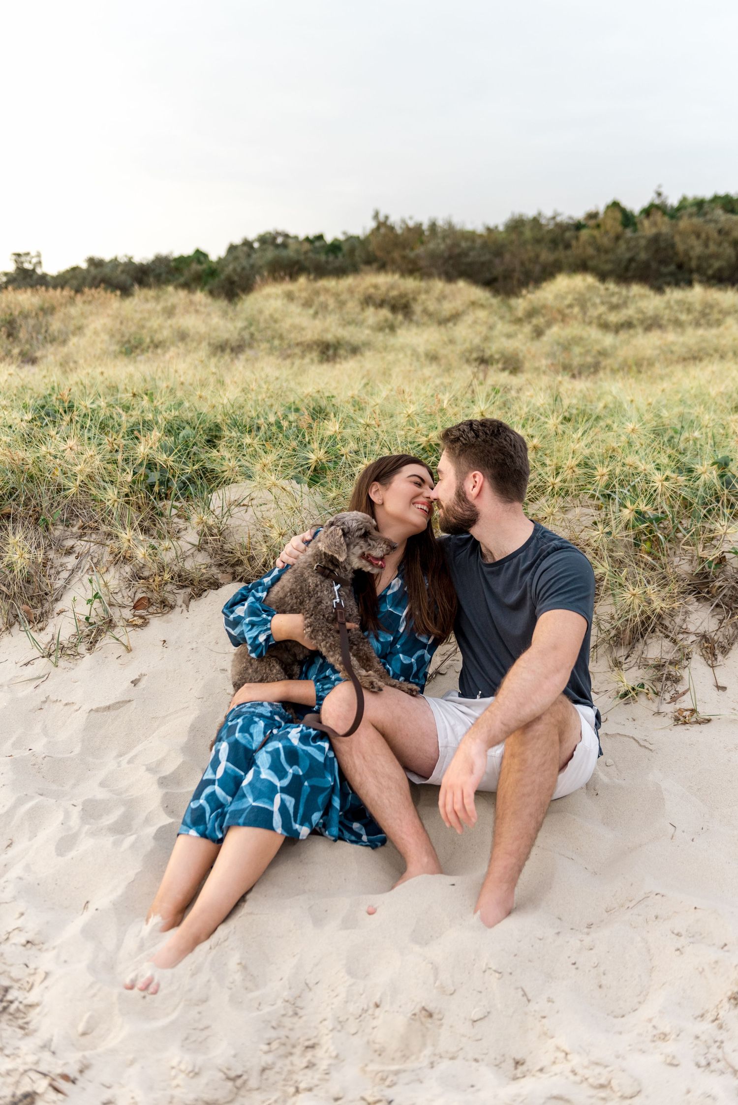 Happy couple sitting on beach sand with gray poodle mix dog climbing on woman in blue dress.