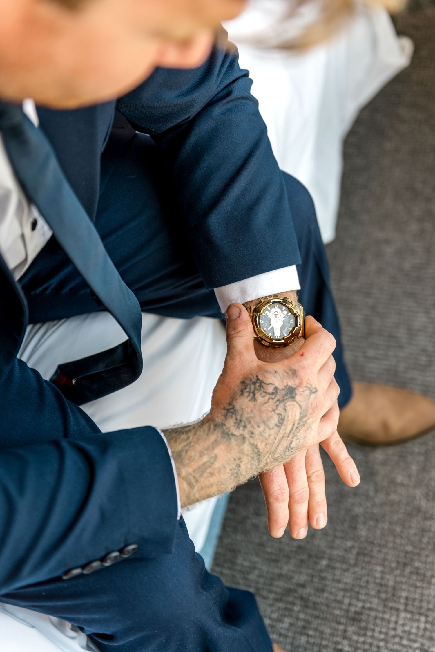 Close-up of groom in navy suit adjusting gold watch on tattooed wrist before wedding.