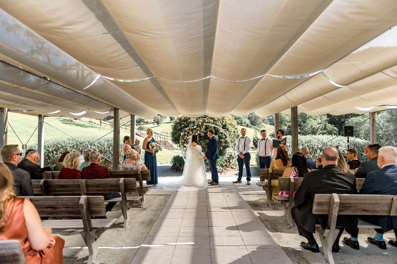 Bride walks down the aisle at an outdoor wedding ceremony under a canopy with floral arch.
