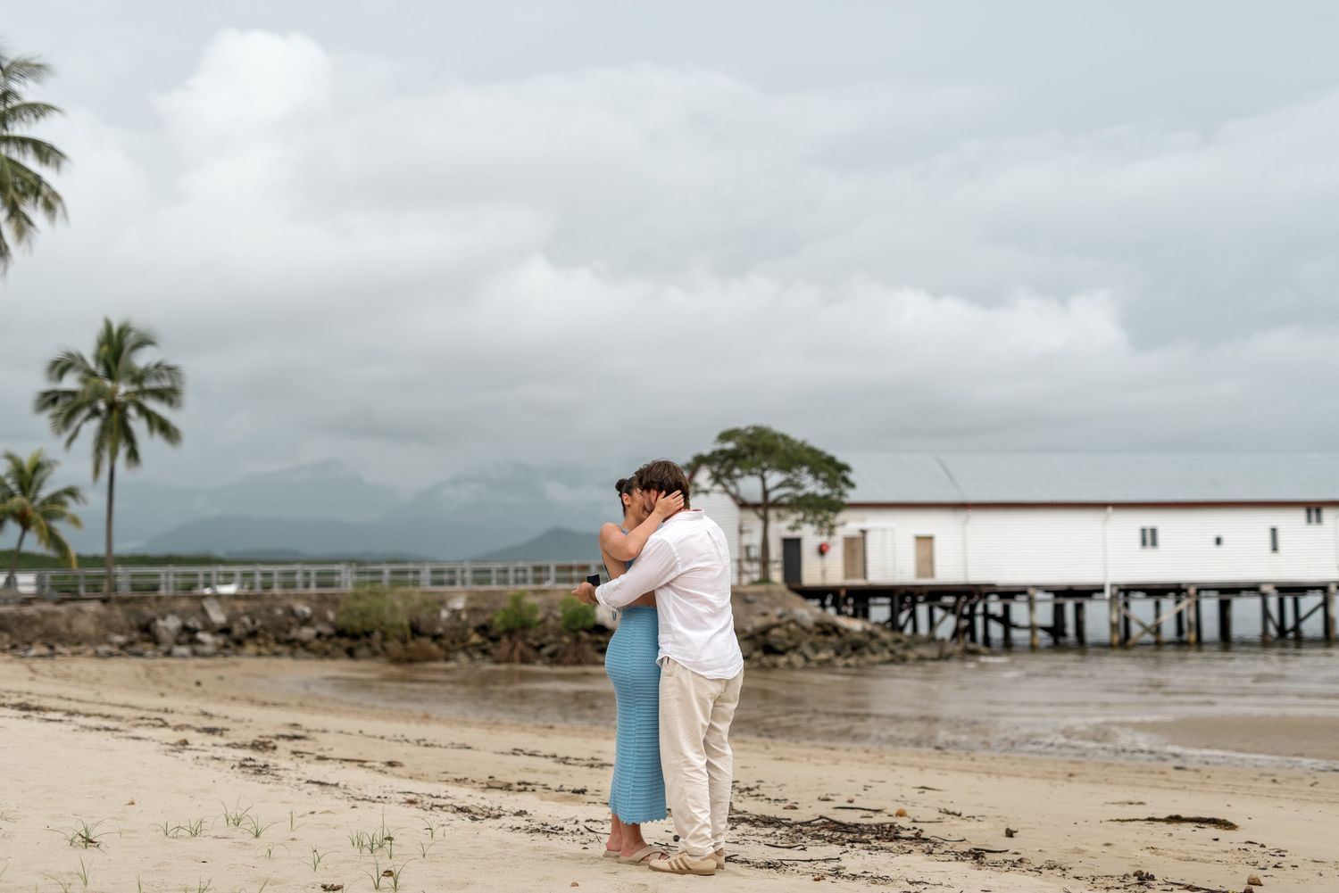 Newly engaged couple embracing on tropical beach with palm trees and pier in background.
