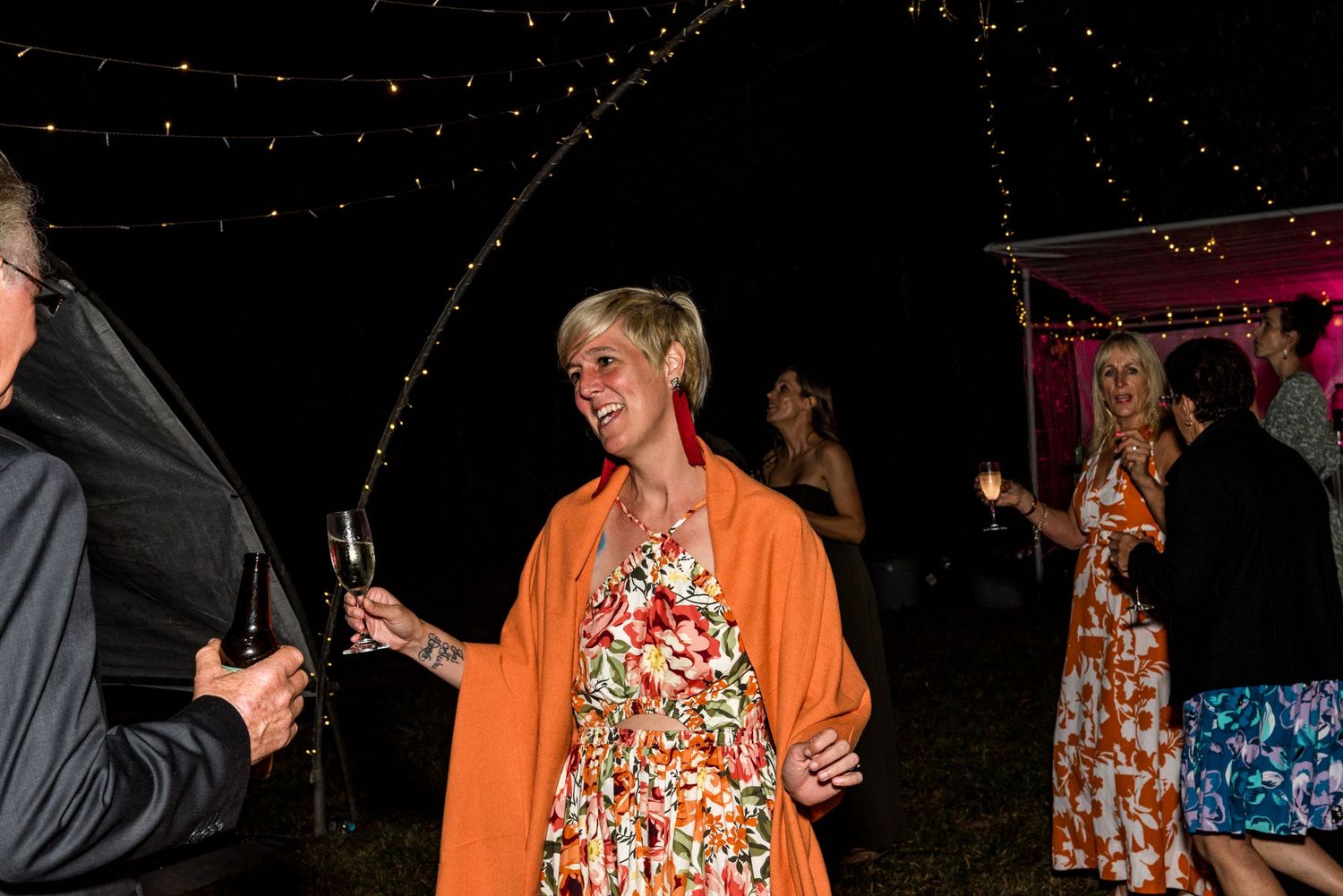 Smiling woman in orange cardigan and floral dress holding champagne glass at outdoor night event.