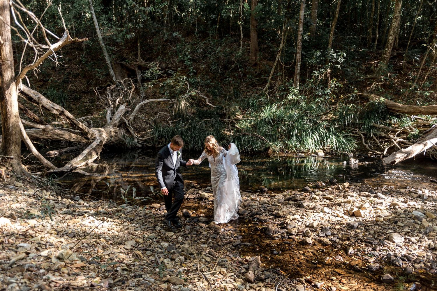 Bride and groom holding hands walking through rocky forest creek bed surrounded by trees.