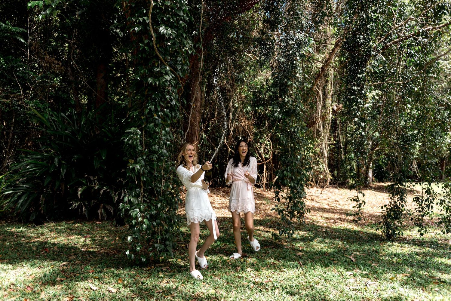 Two women in white dresses playing on a swing surrounded by tall green trees outdoors.