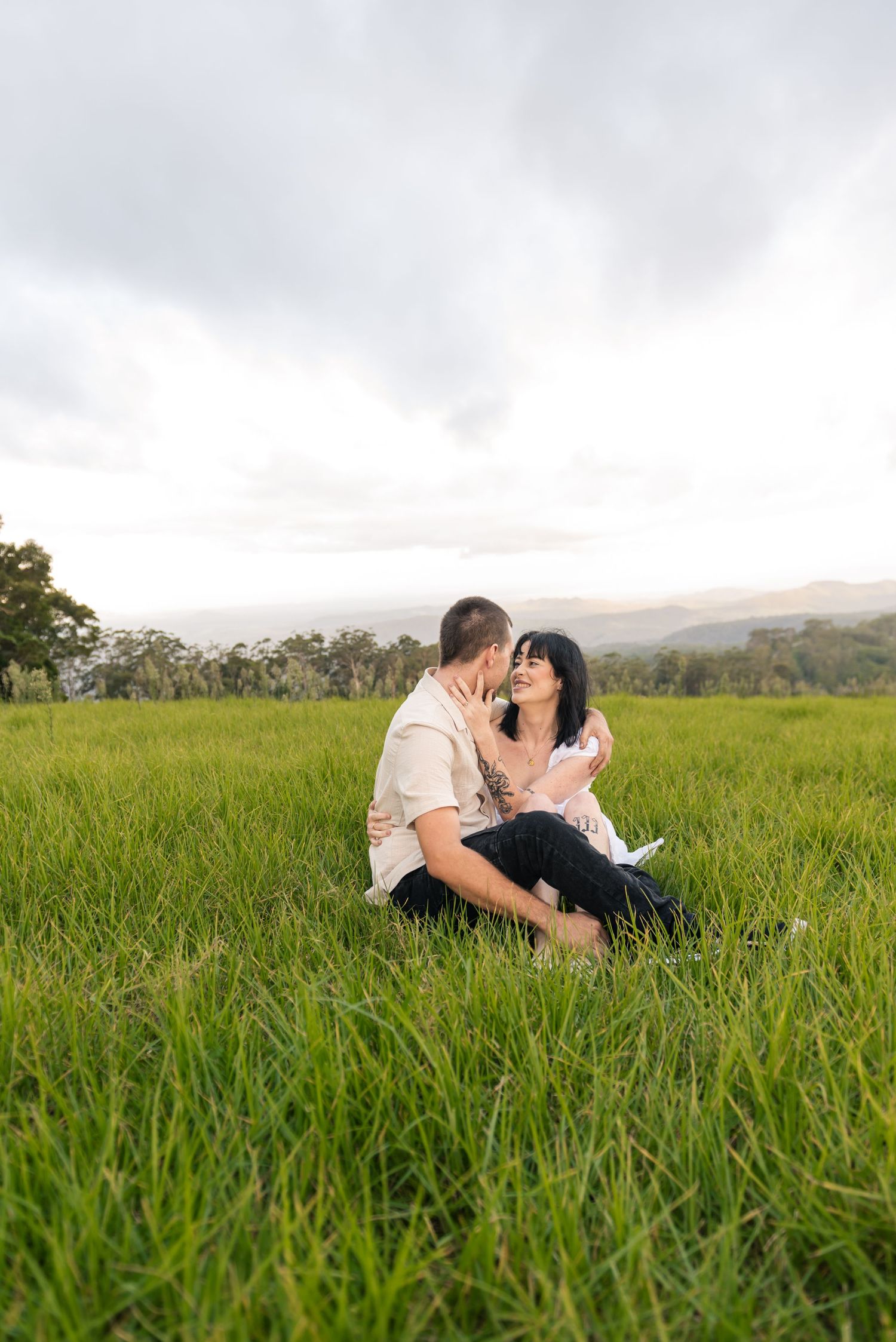 Smiling couple sitting closely together in a lush green meadow under cloudy skies.