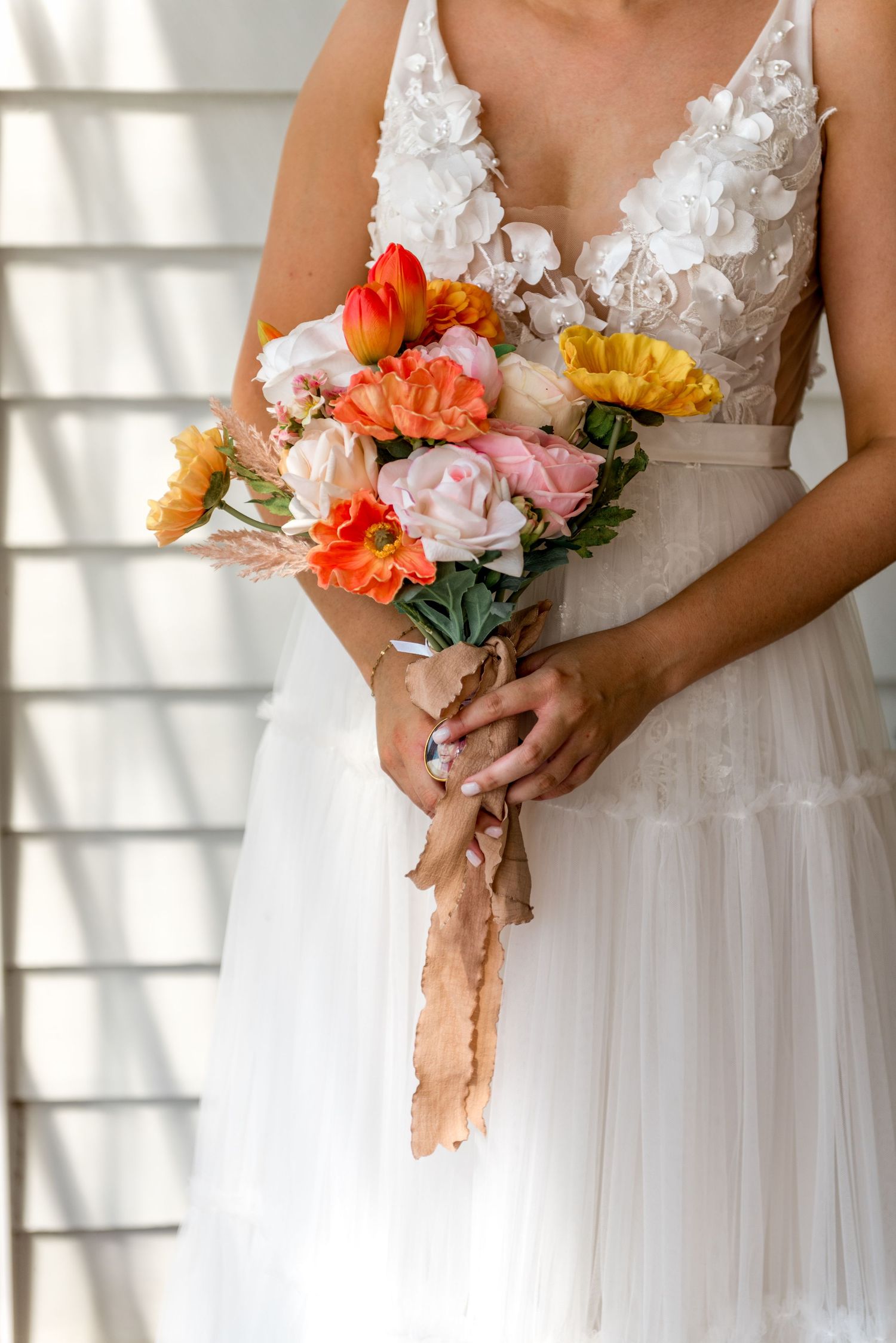 Bride holding vibrant orange, pink and yellow floral bouquet wrapped in tan ribbon against white dress.