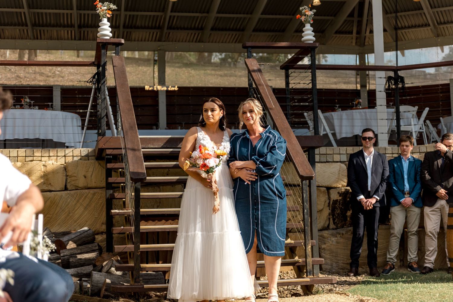 Bride in white gown walking down the aisle with escort at a rustic outdoor wedding ceremony.