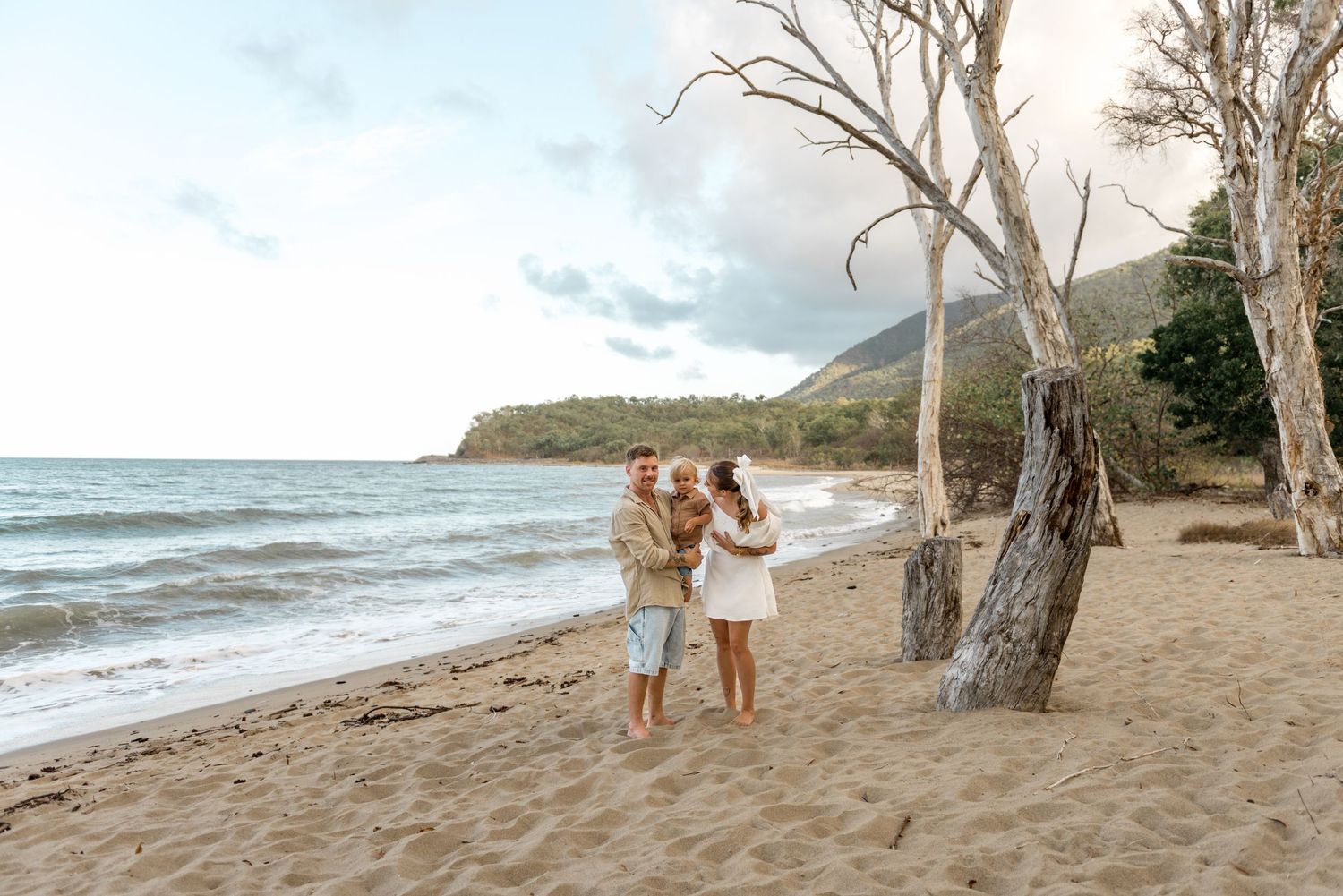 Family of three standing together on wide sandy beach with calm ocean and hills behind.