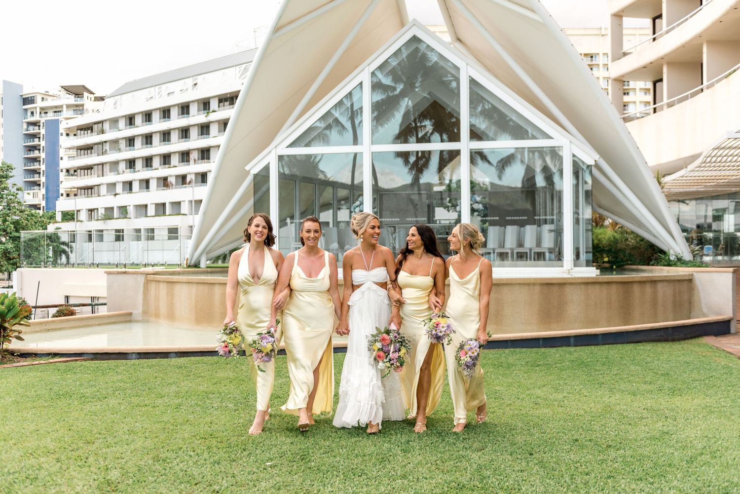 Bride and four bridesmaids in cream dresses pose on lawn before a geometric glass wedding venue.