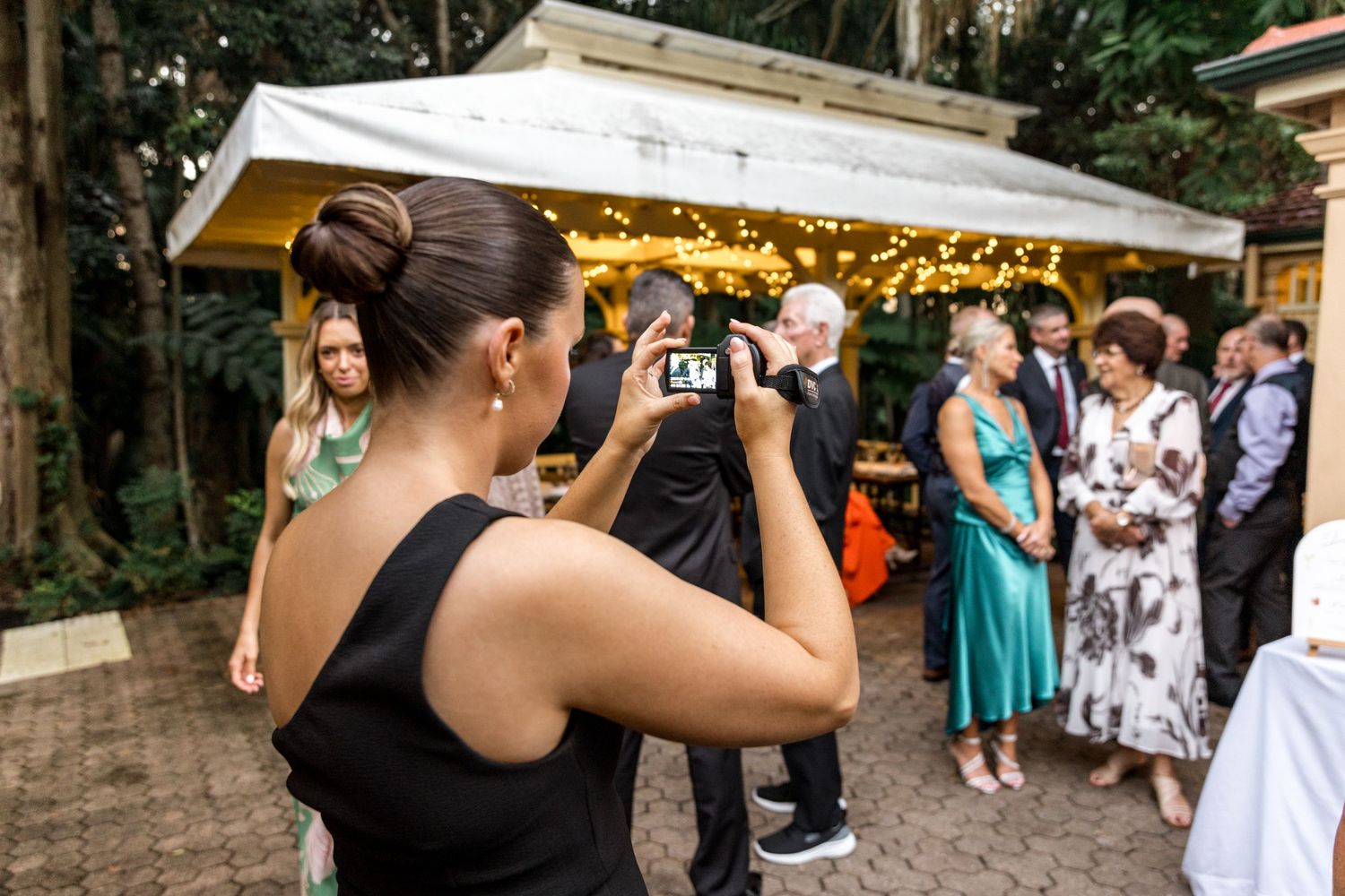 Wedding guest photographs a group of elegantly dressed attendees at an outdoor reception with fairy light canopy.