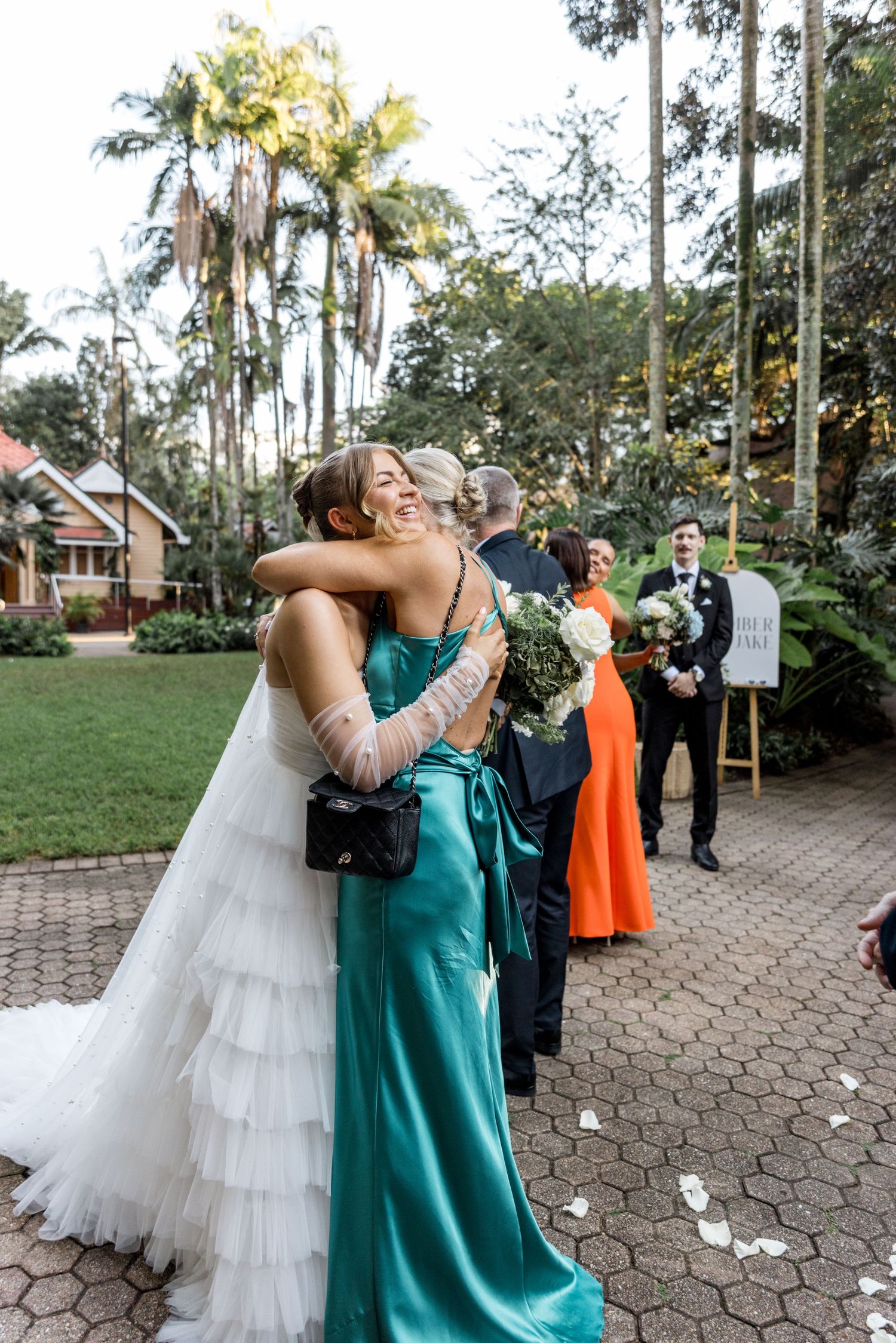 Bride in ruffled white gown embraces a guest in teal satin dress during outdoor garden wedding celebration.