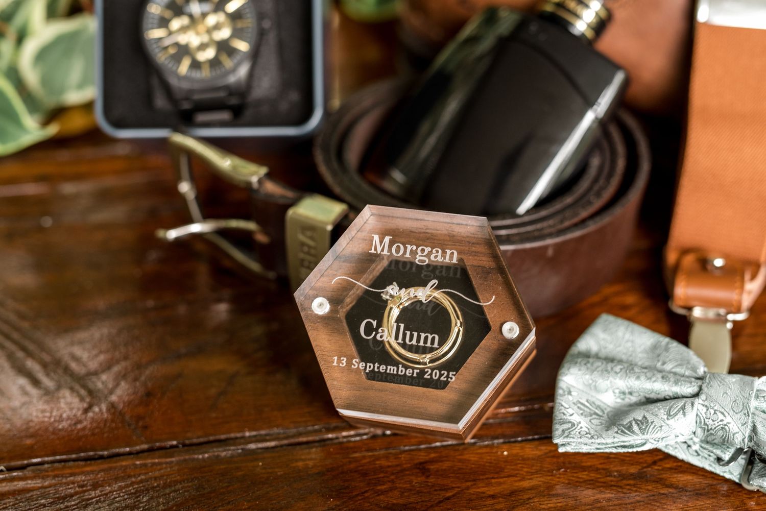 Personalized hexagonal wooden ring box engraved with Morgan and Callum wedding date beside a flask and leather belt.