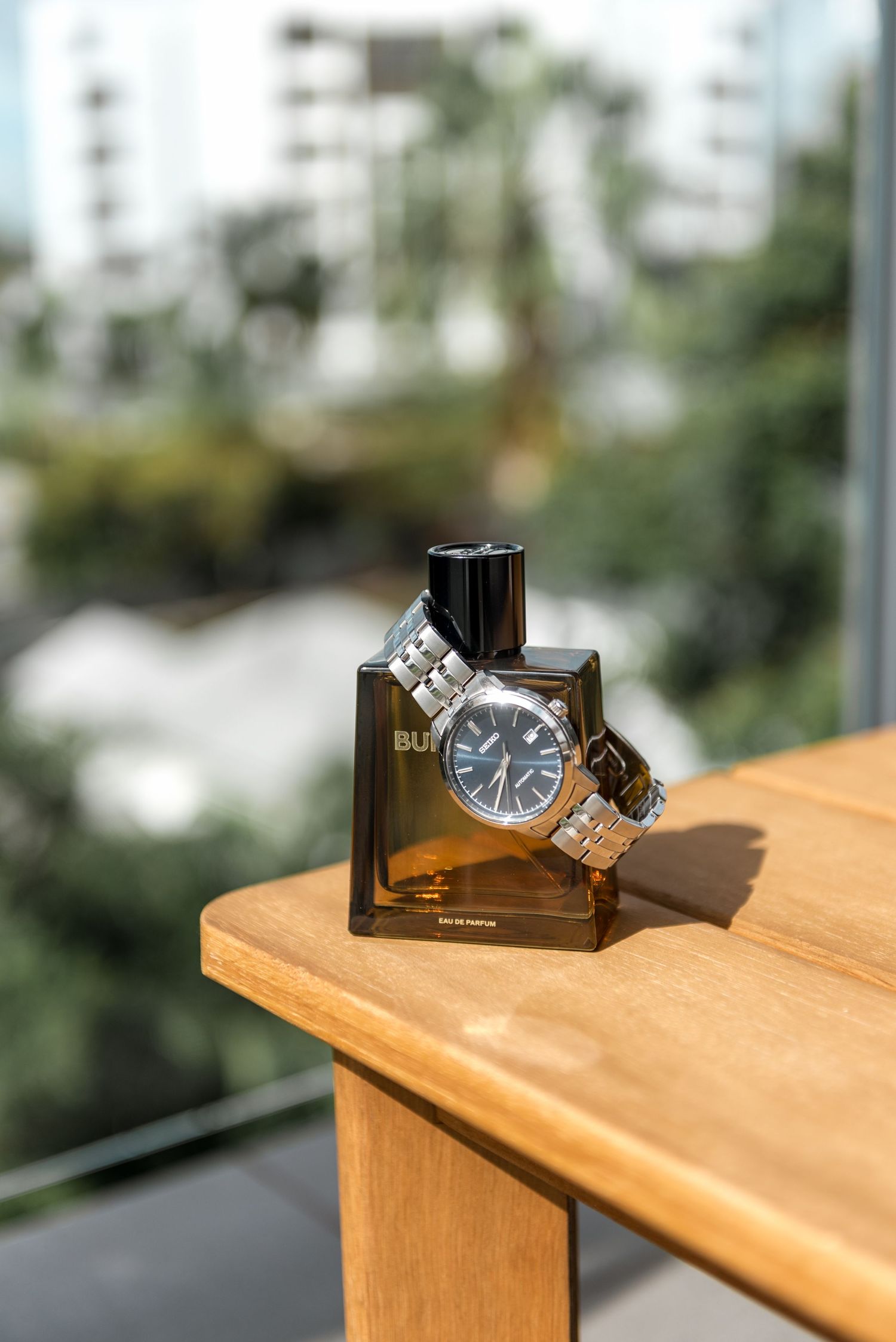 Burberry cologne bottle with silver watch on wooden table with city view background.