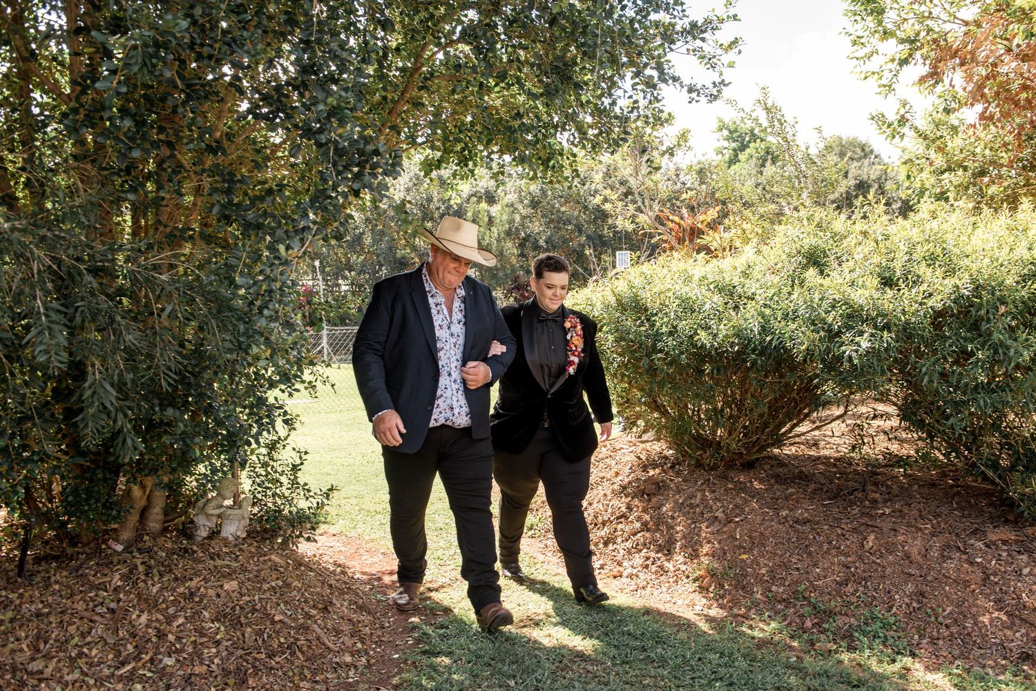 Two men in suits walk together through a lush green garden path on a sunny wedding day.