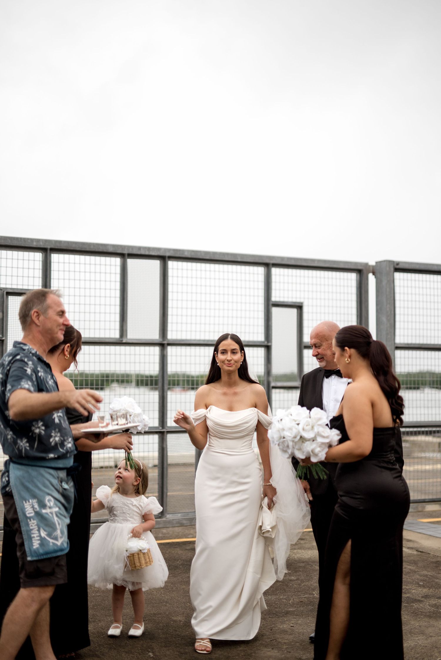 Bride in white gown with wedding party gathered near waterfront security fence.