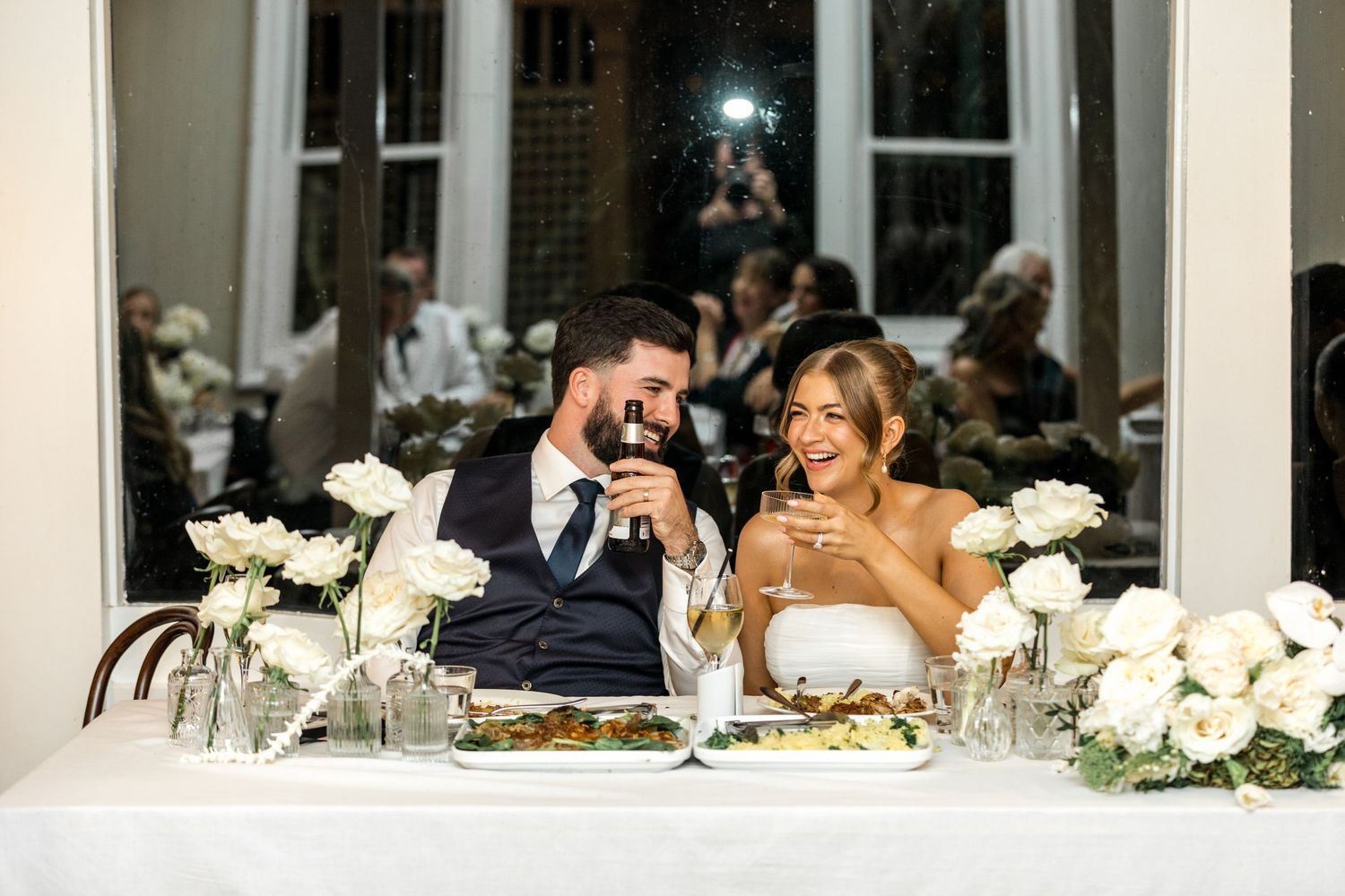 Bride and groom smiling and toasting with wine glasses at their wedding sweetheart table.