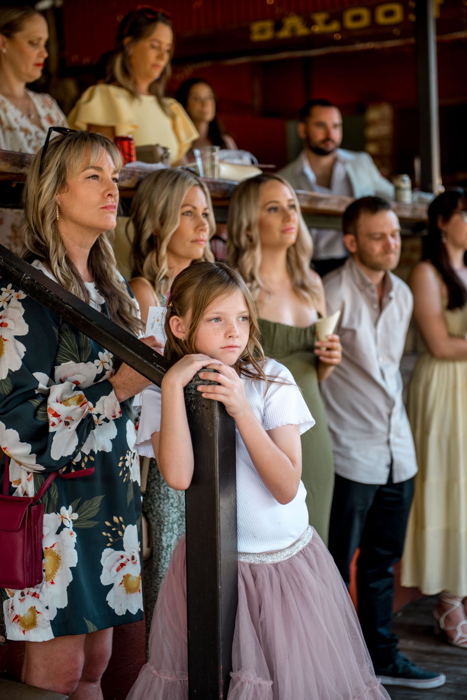 Wedding guests including young girl in pink tulle skirt watch ceremony attentively indoors.