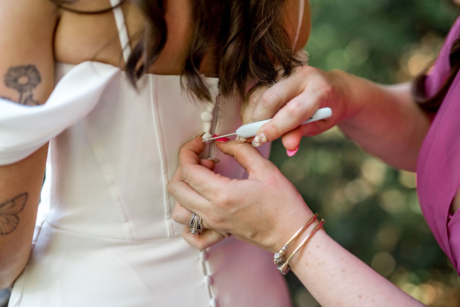 Close-up of bridesmaid fastening buttons on bride's white wedding dress, showing tattoo and jewelry details.