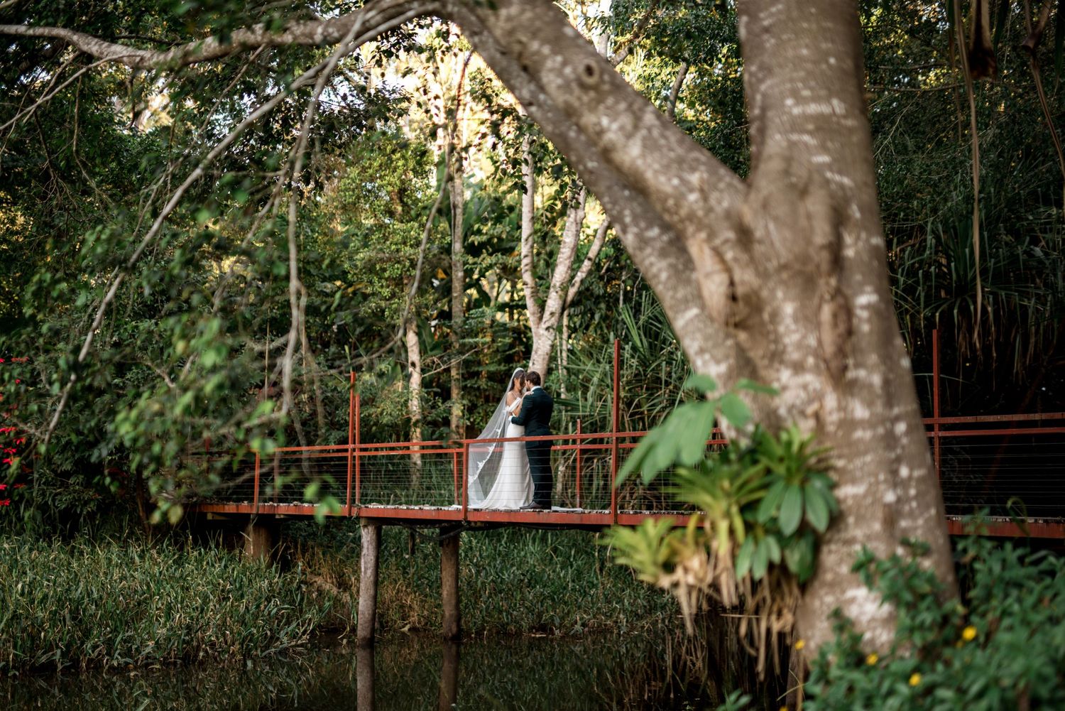 Bride and groom share a romantic moment on a red bridge surrounded by lush greenery.