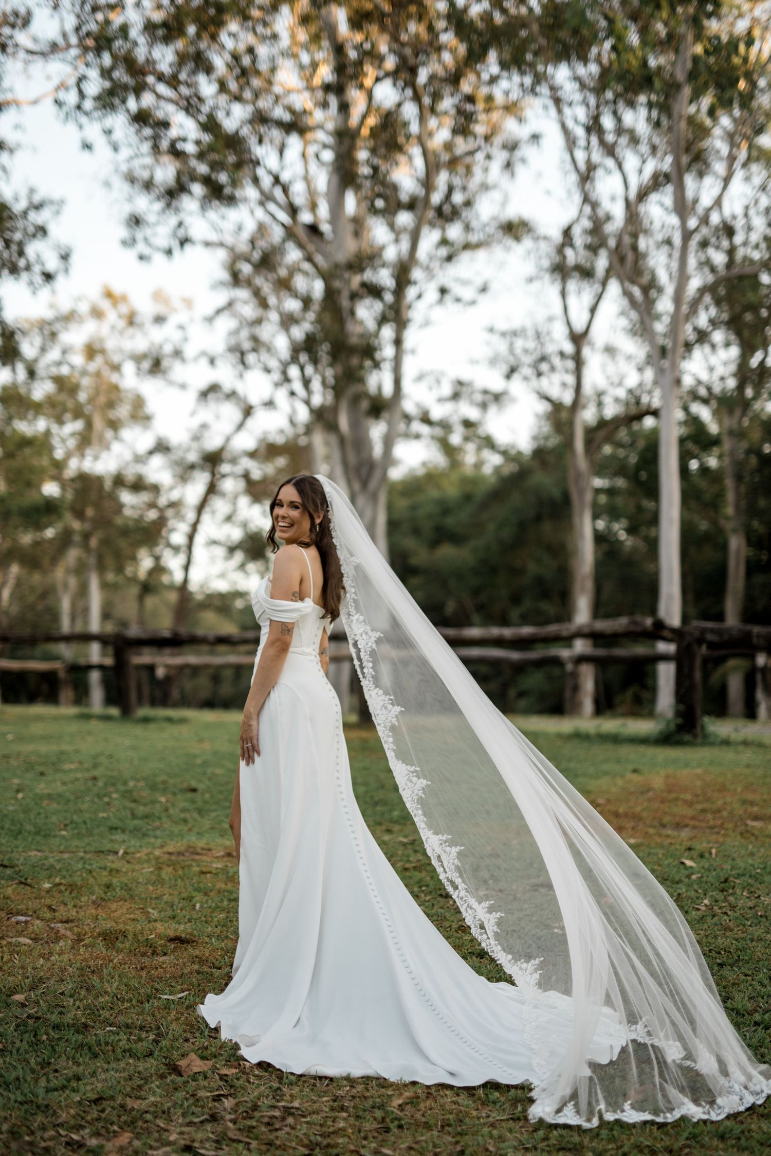 Bride in off-shoulder gown with flowing lace veil posing in a rustic outdoor setting.