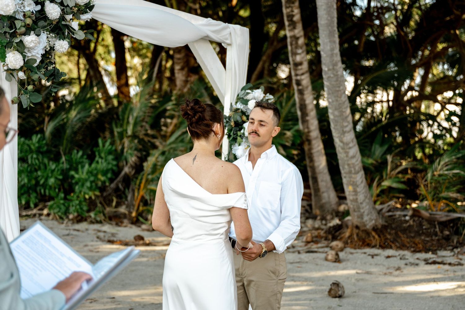 Bride and groom exchanging vows at beach wedding altar, officiant reading from book, tropical palm trees behind at Four Mile Beach.