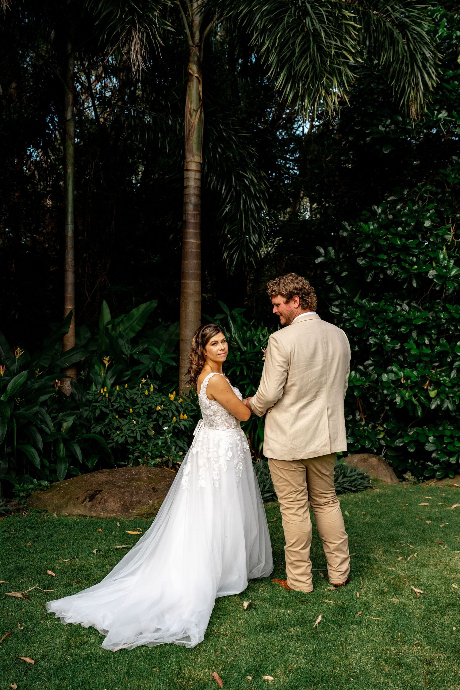 Bride glances back while holding groom's hand near tropical palm trees.