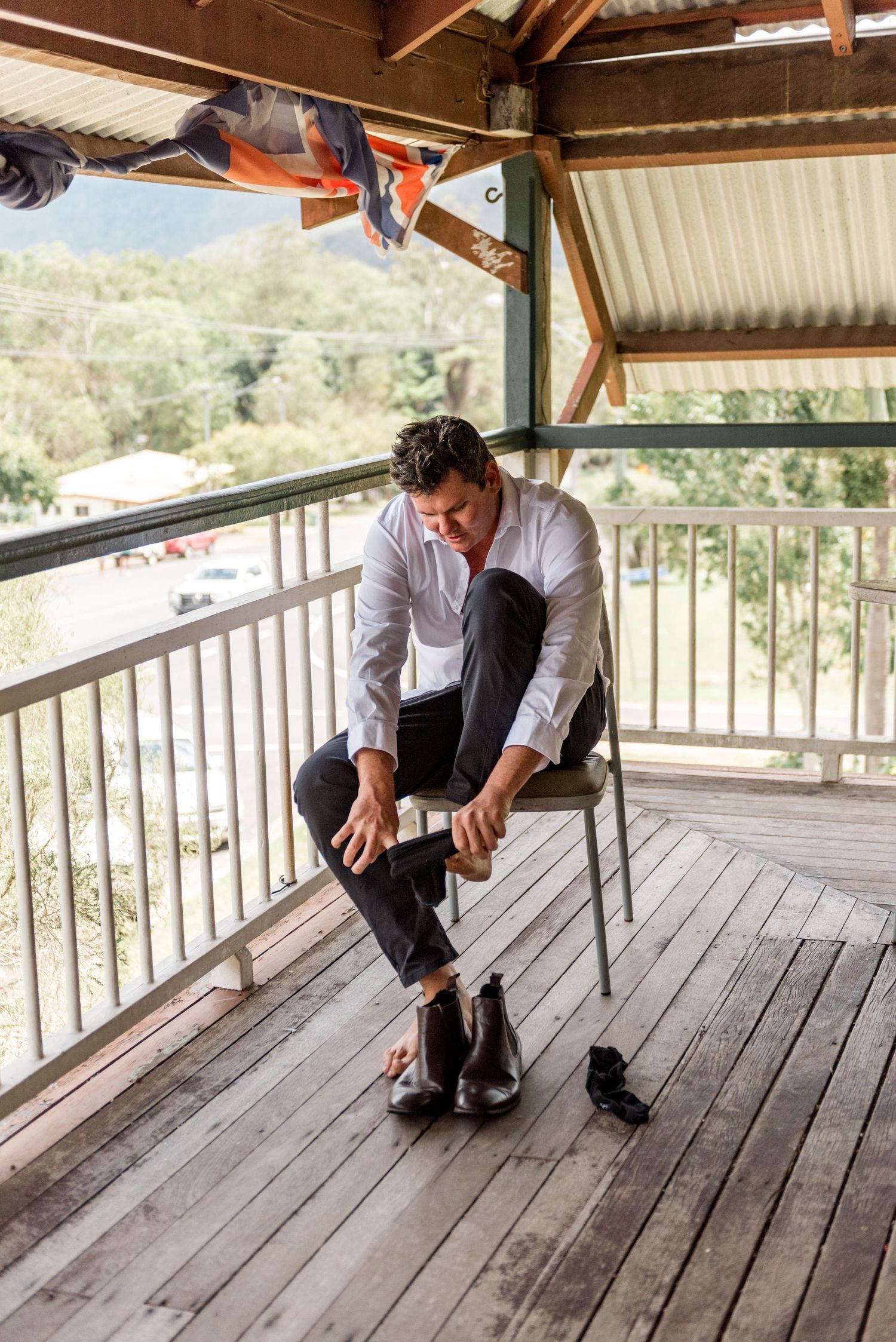Groom in white shirt putting on brown leather boots while seated on a rustic outdoor porch.