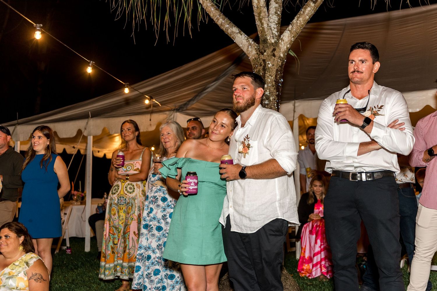 Wedding guests laughing and celebrating outdoors at night under string lights near white marquee tent at reception.
