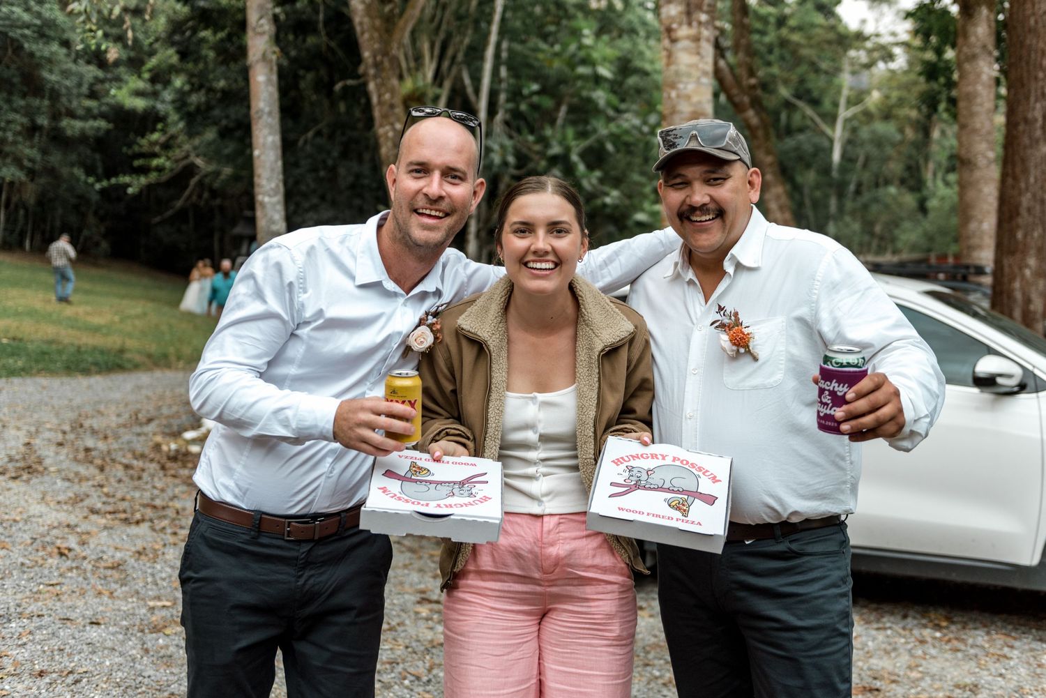 Two men and young woman smiling together holding drinks and pizza boxes at outdoor celebration.