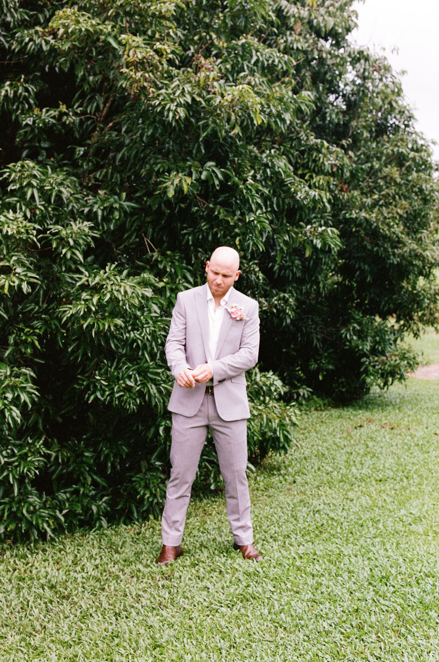 Groom in light gray suit adjusting cufflinks outdoors on green lawn with lush trees behind him.