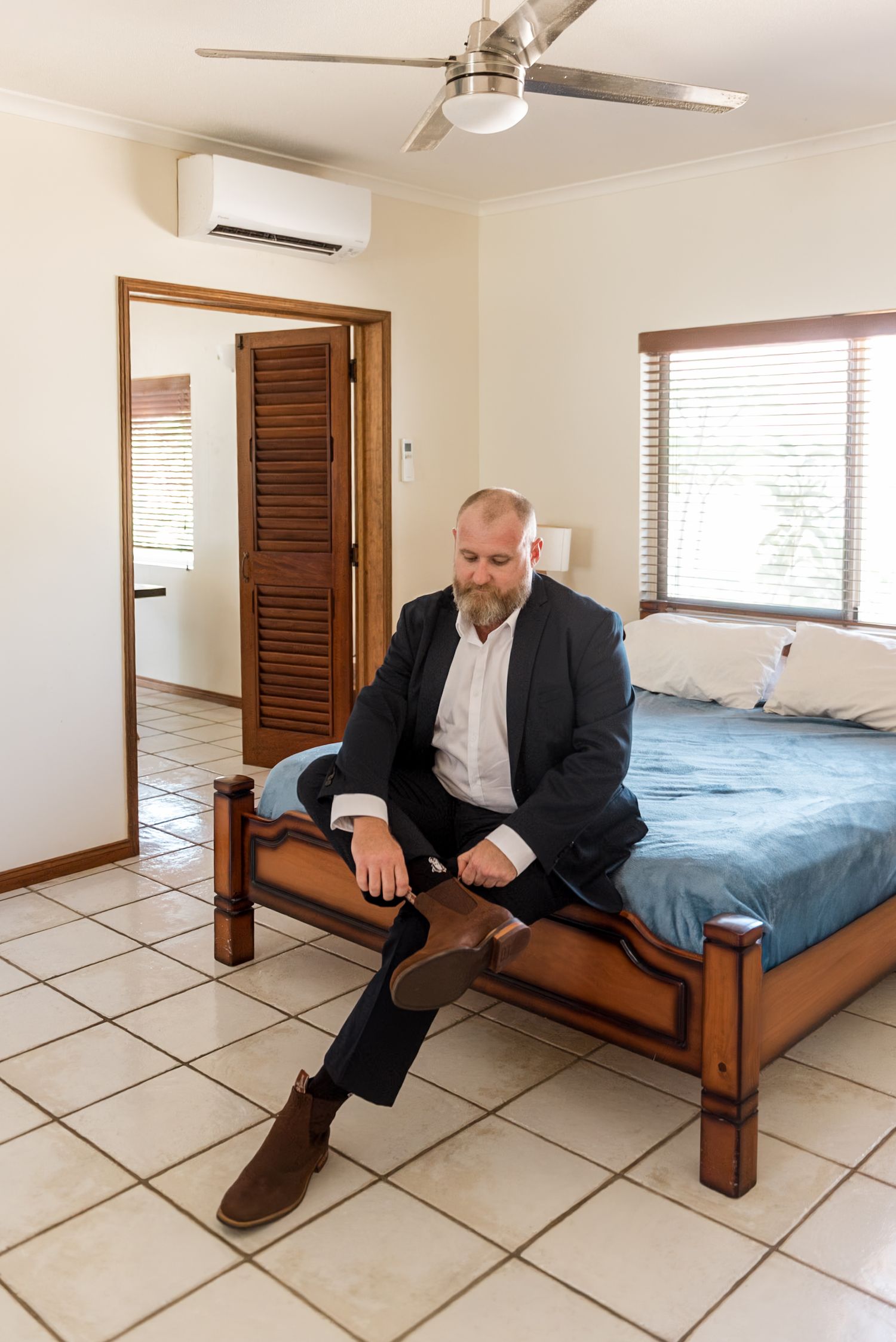 Bearded groom in navy suit sitting on bed putting on brown boots in bright bedroom.