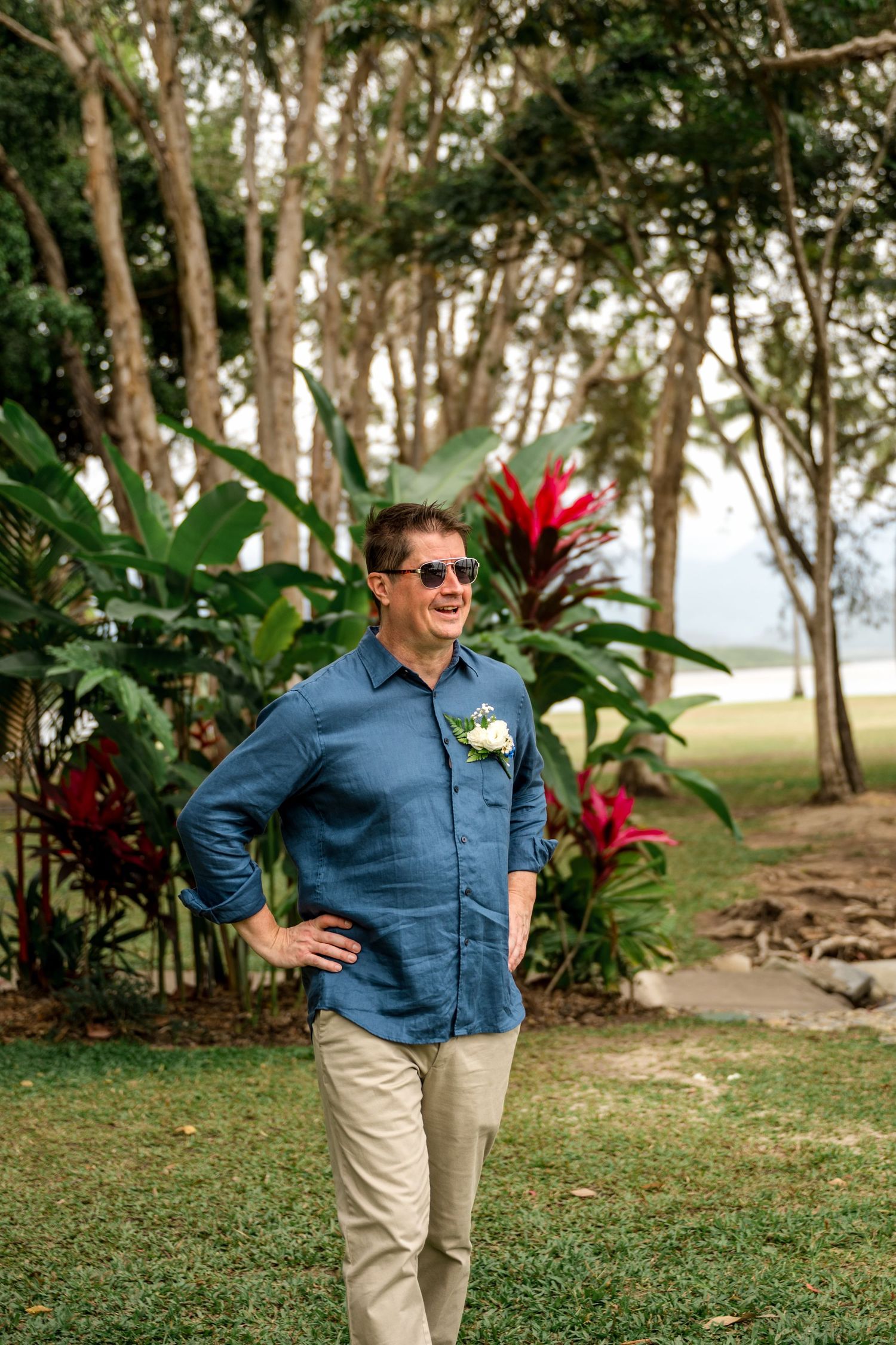Smiling groom in blue linen shirt with boutonniere stands in tropical garden setting at Little Cove, Port Douglas.