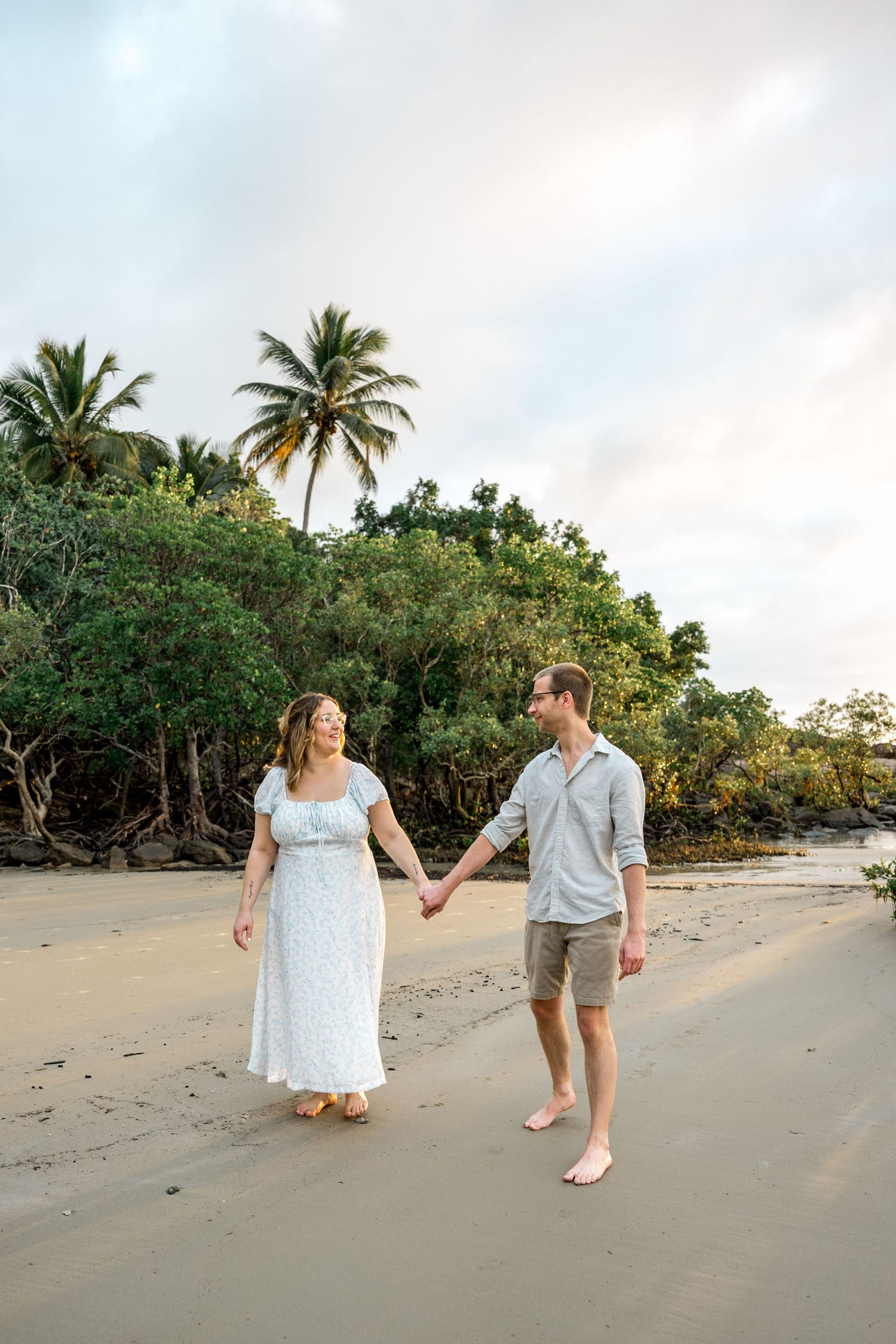 Couple holding hands and smiling while walking barefoot on sandy beach with palm trees at sunset at Four Mile Beach, Port Douglas.