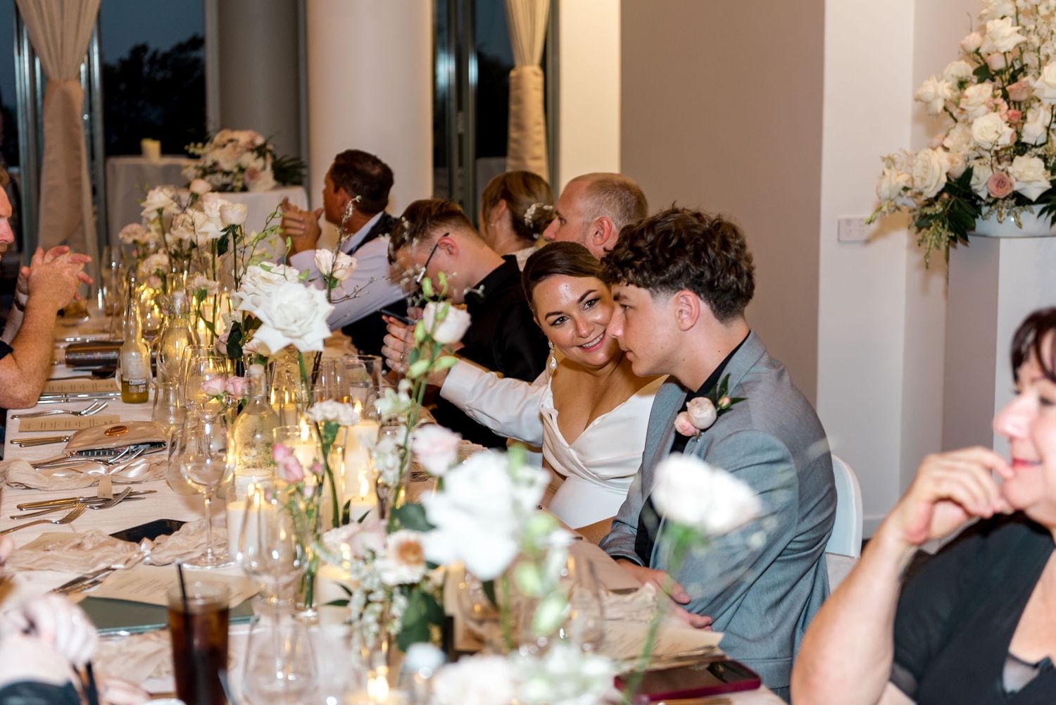 Bride and groom lean heads together affectionately at long decorated wedding reception dinner table.