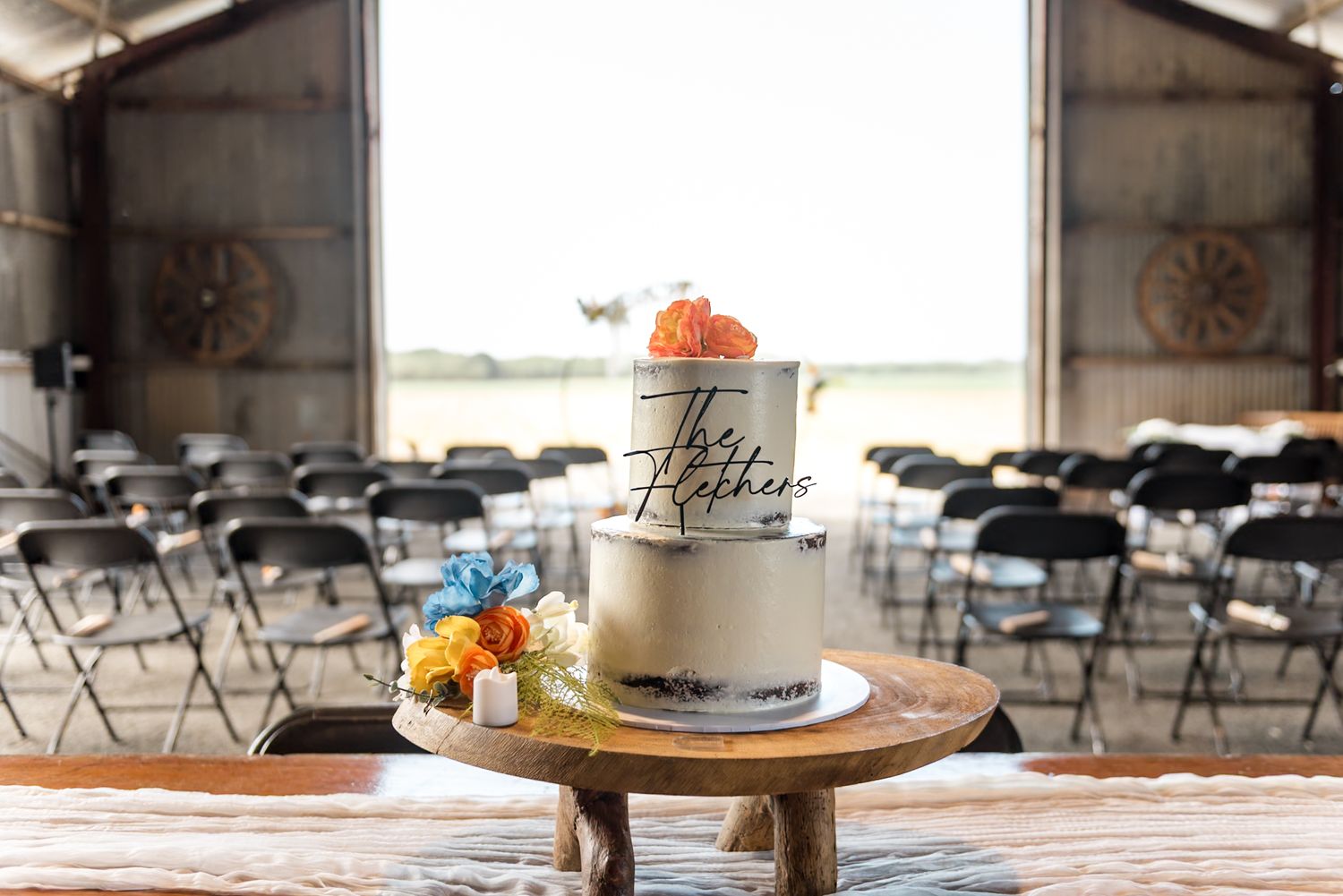 Two-tier semi-naked The Fletchers wedding cake on wooden stand in rustic barn ceremony venue.