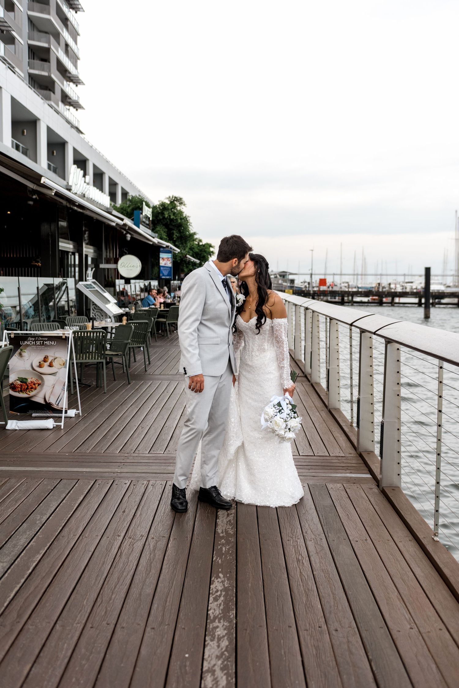 Newlyweds share a kiss on a waterfront boardwalk, bride holding a white bouquet near a marina.