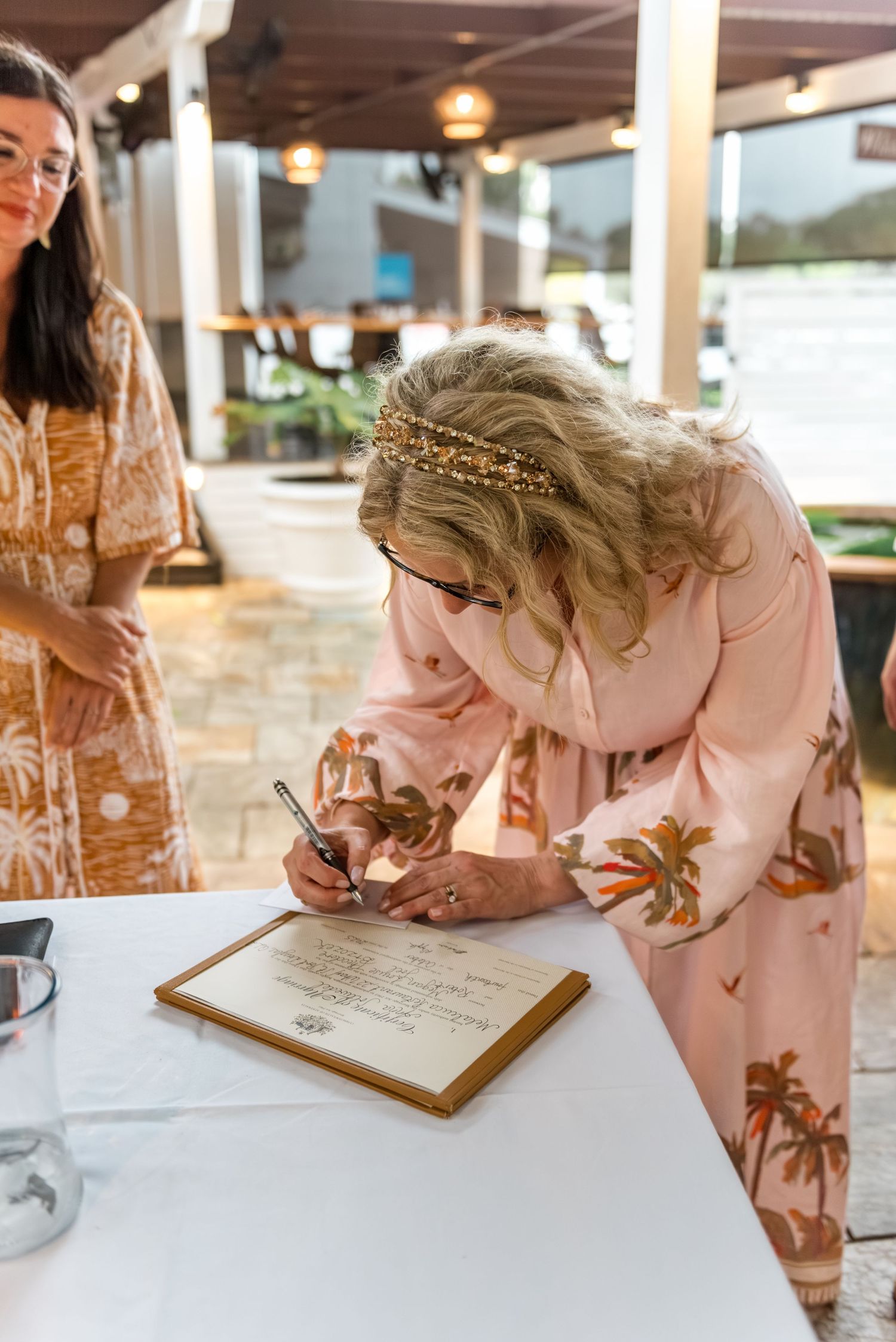 Bride wearing floral crown signing wedding certificate at outdoor ceremony table with witness standing nearby.