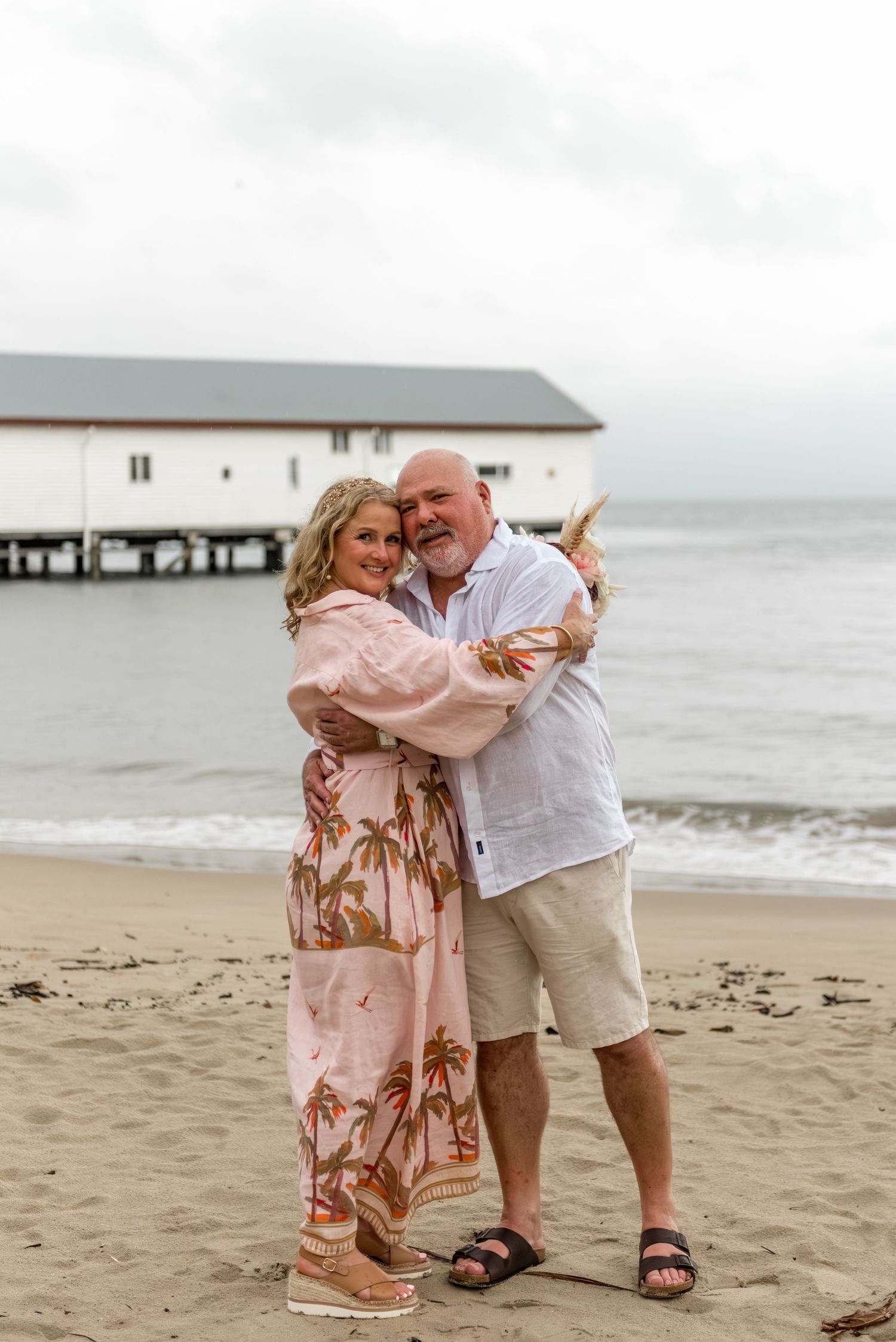 Couple embracing warmly on a sandy beach with a white boathouse pier in the background.