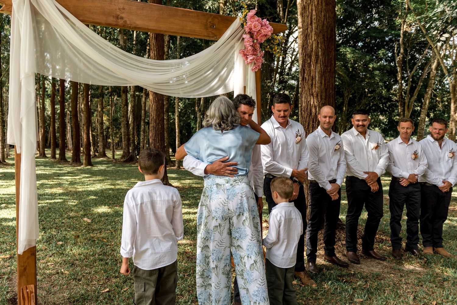 Groom embraces a guest near the floral wedding arch surrounded by groomsmen in a forest.
