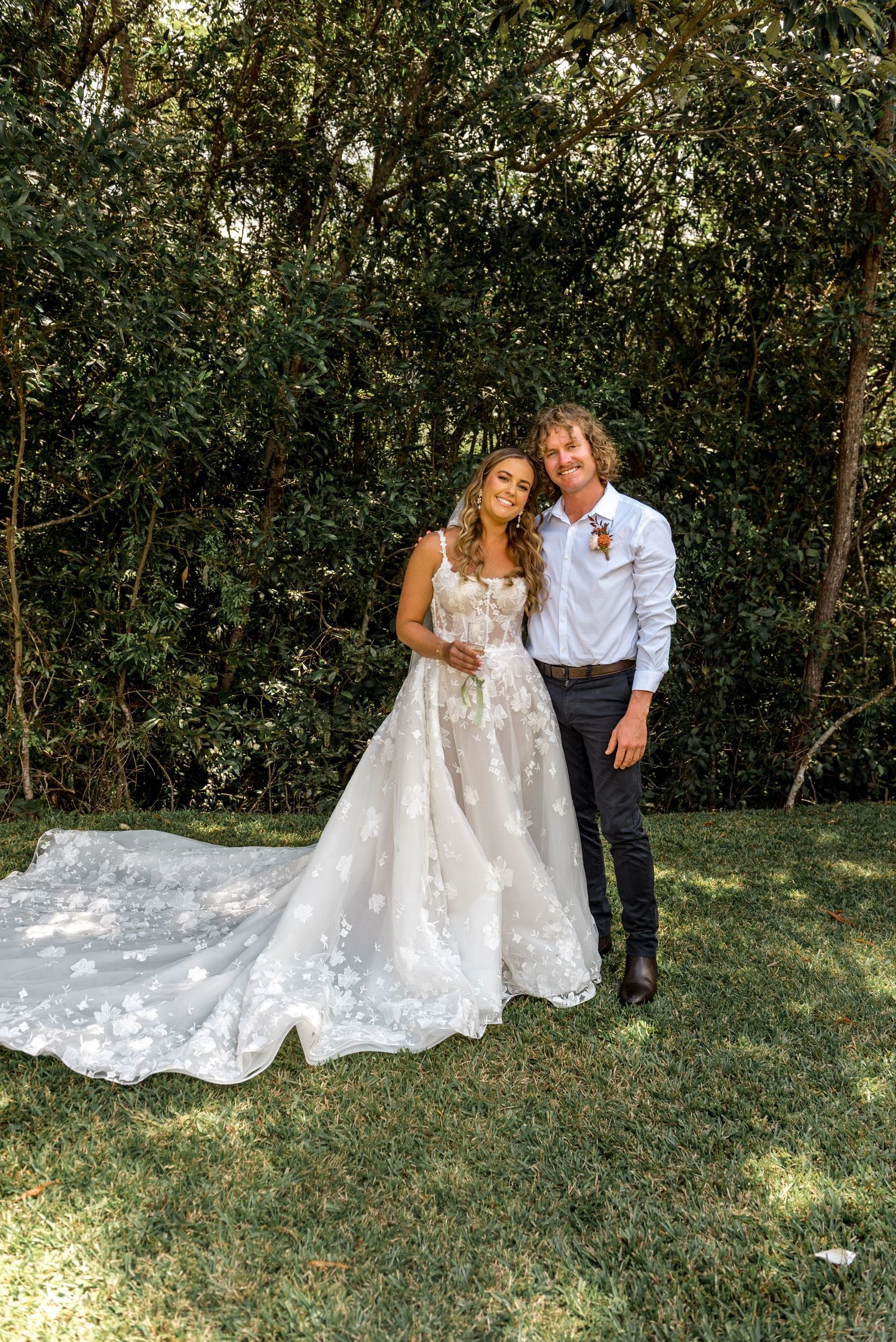 Smiling bride and groom pose together in garden setting with lush green trees behind them.