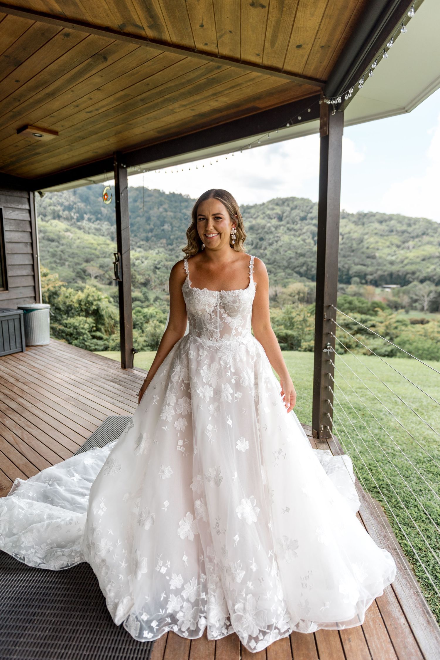 Smiling bride in floral lace ball gown standing on wooden deck with lush green mountain backdrop.