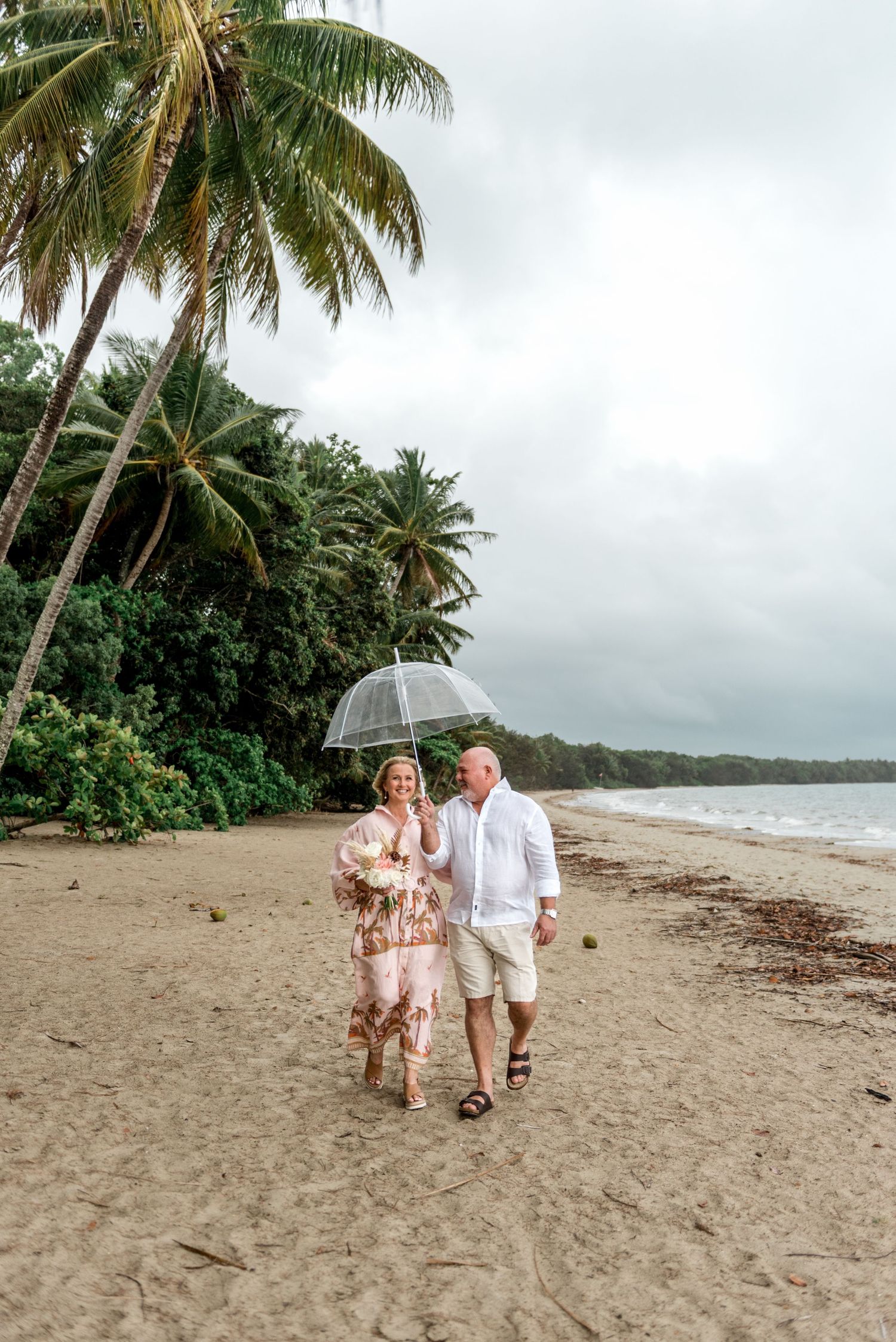Smiling couple walking hand in hand on a tropical beach under a clear umbrella together.