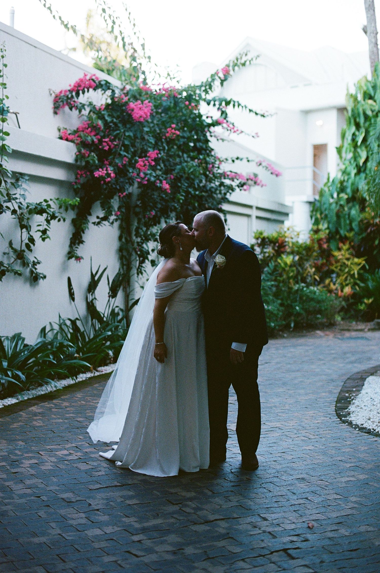 Bride and groom sharing a romantic kiss on a brick pathway surrounded by lush tropical gardens and pink bougainvillea.