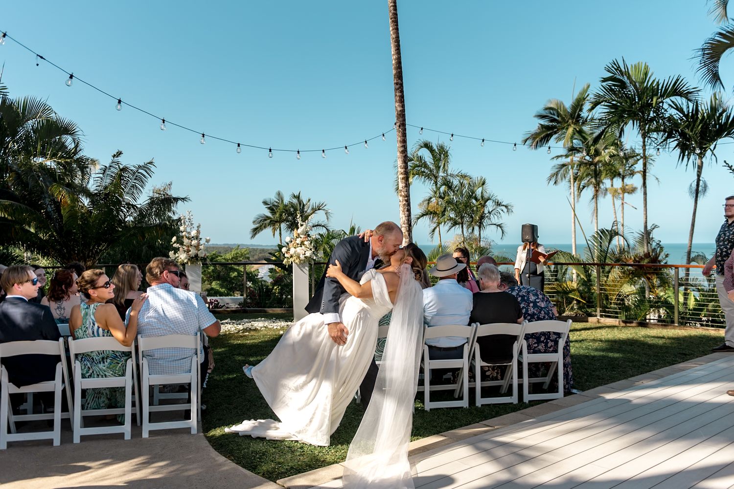 Newlyweds share romantic first kiss at outdoor tropical wedding ceremony with guests seated in white chairs.