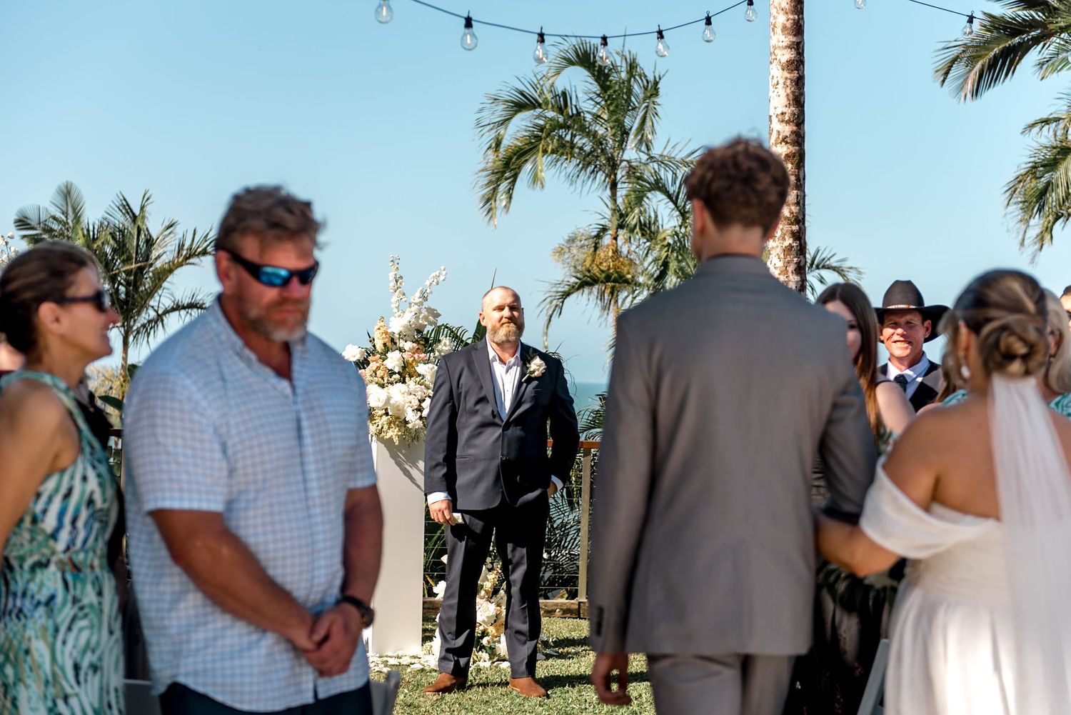 Groom awaits bride at outdoor tropical wedding ceremony with palm trees and string lights overhead.