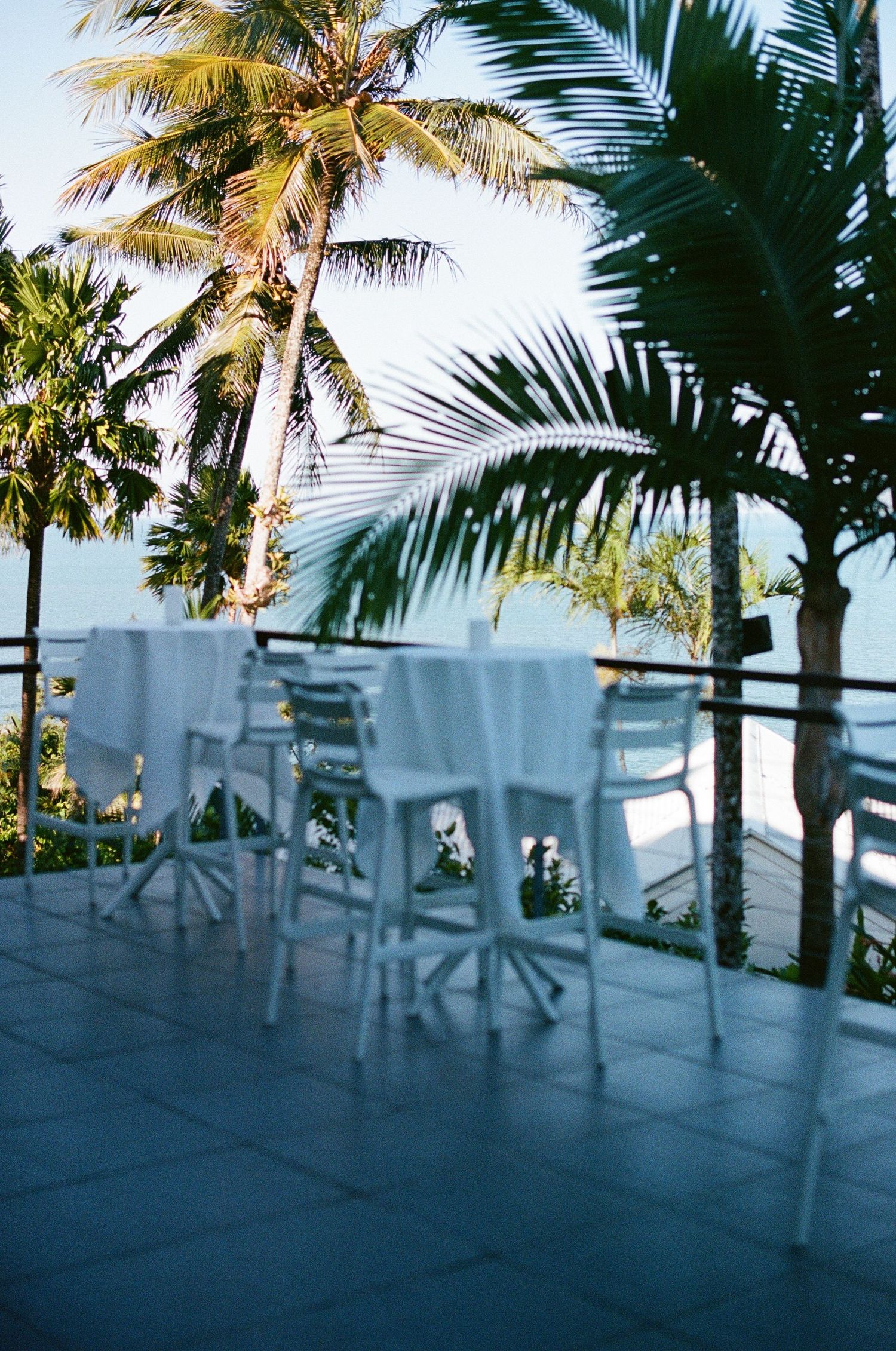 Elegant white chairs and tables on a tropical outdoor terrace overlooking palm trees and ocean view.