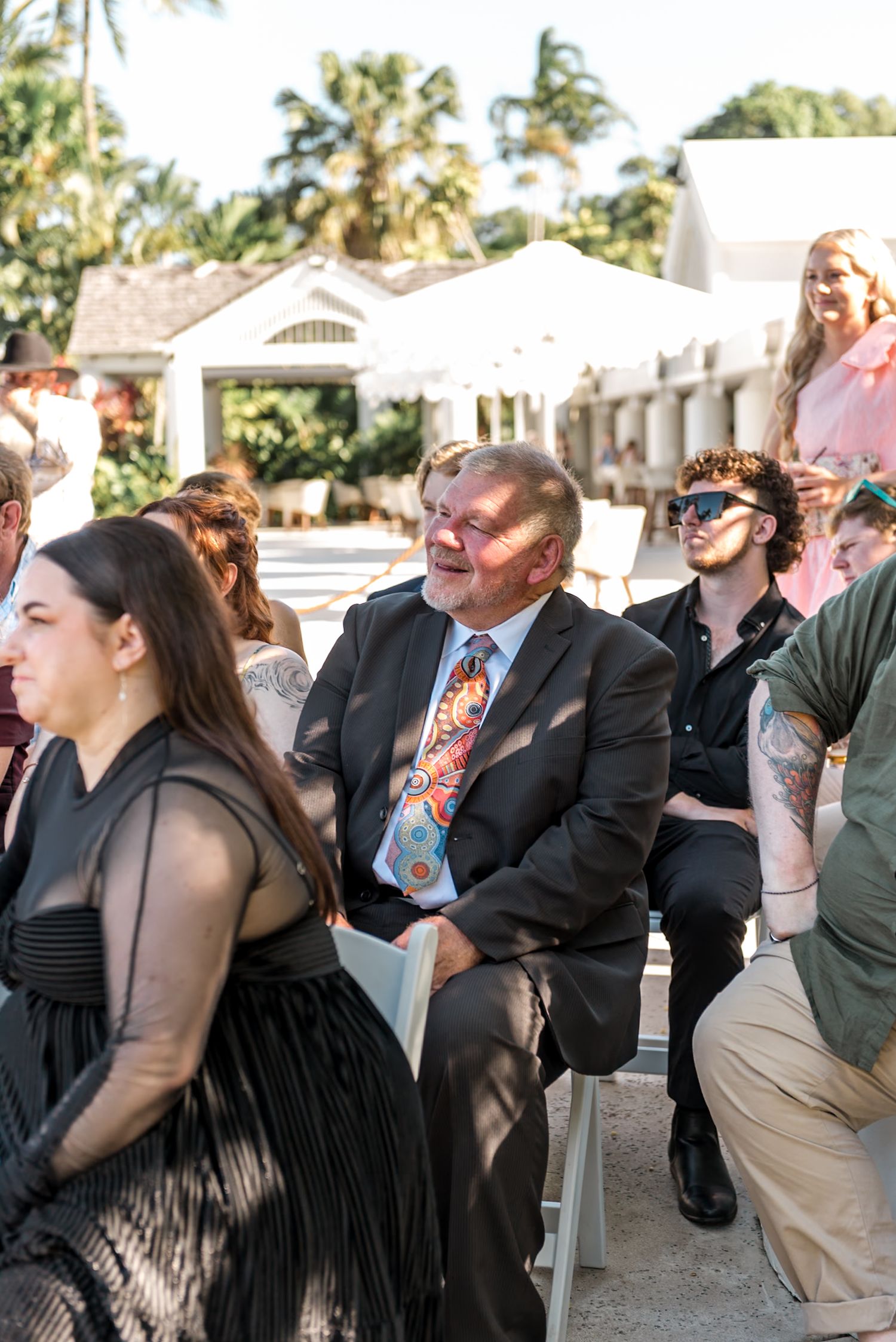 Smiling wedding guest in colorful tie laughs joyfully while seated among guests at outdoor tropical ceremony.