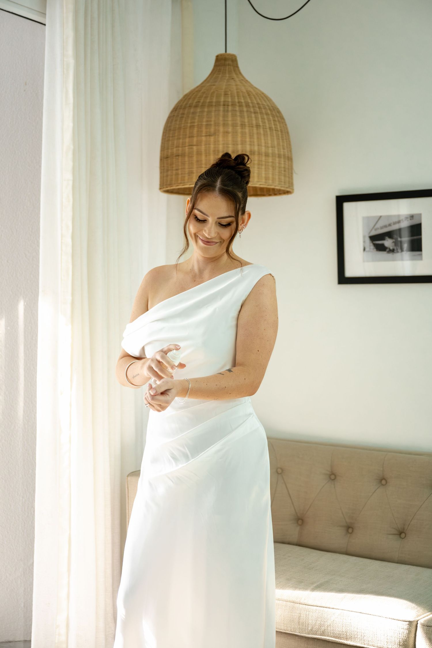 Smiling bride in one-shoulder white satin gown adjusts bracelet in sunlit room with rattan pendant light.