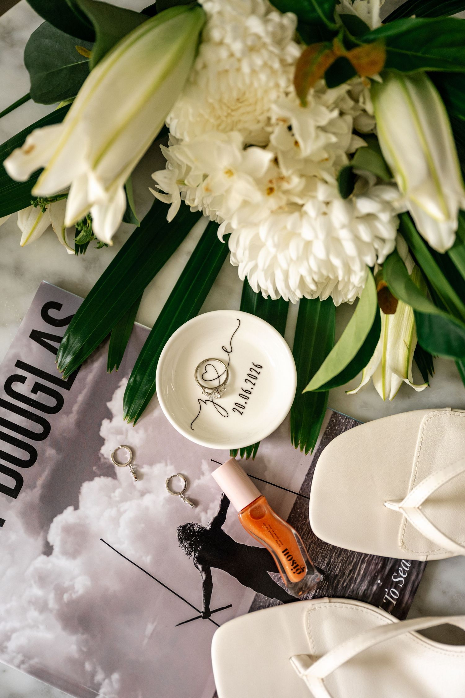 Close-up of personalized wedding ring dish with diamond ring beside white lilies and bridal sandals.