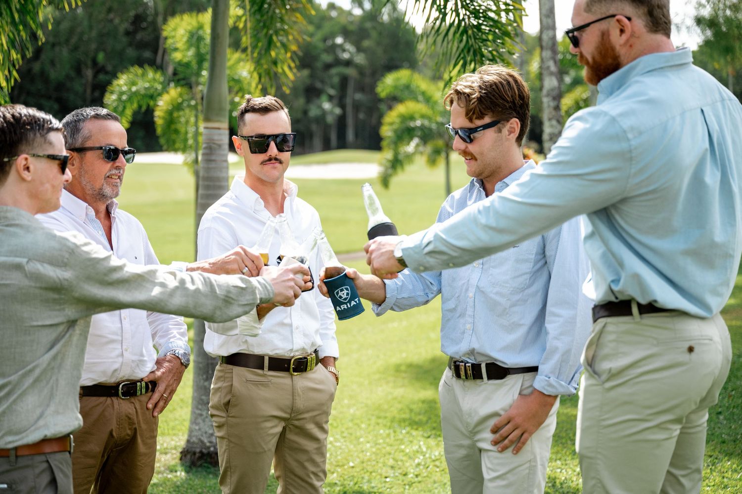 Group of five men in sunglasses toasting drinks together on a sunny tropical golf course.